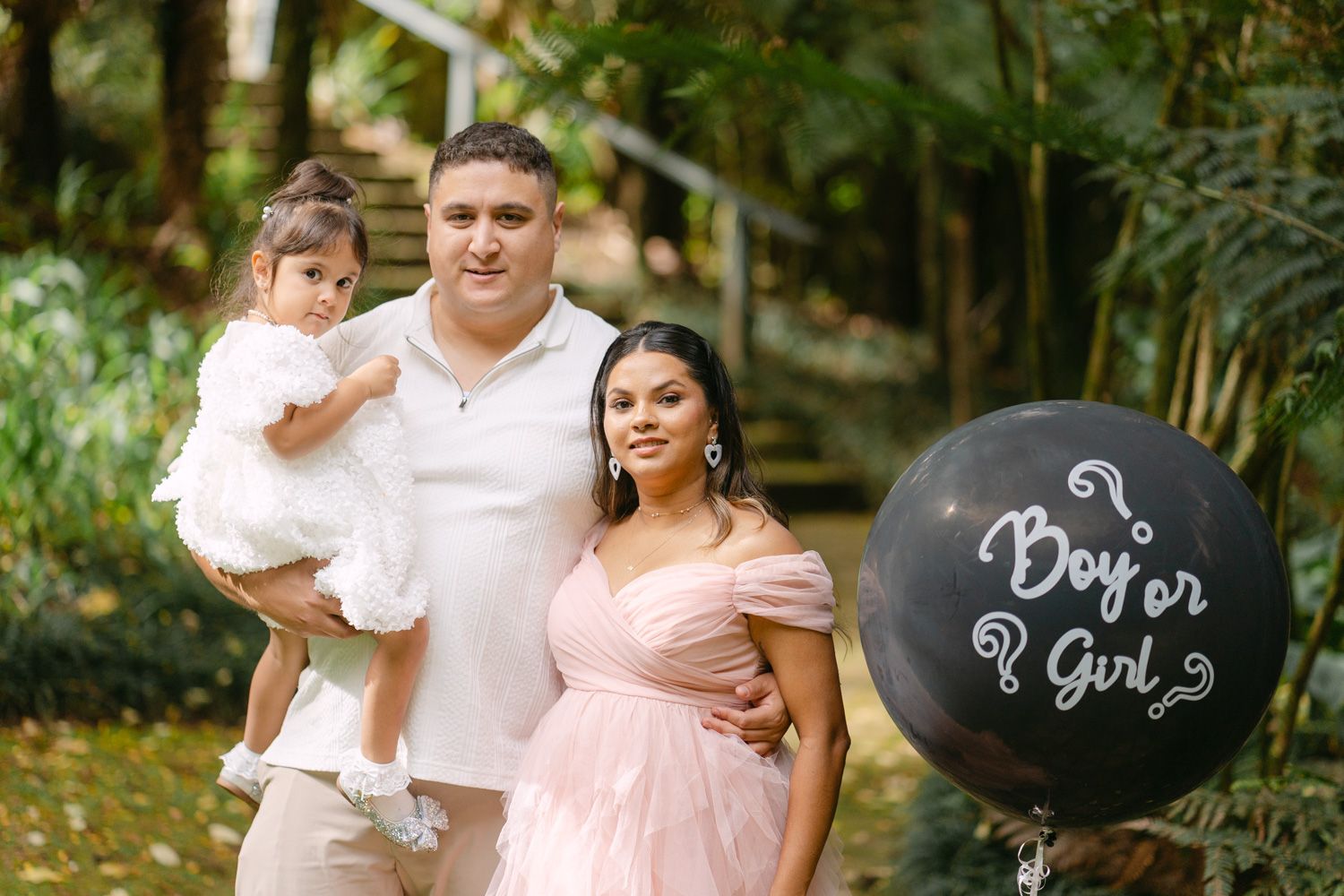 Family poses outdoors at gender reveal with black Boy or Girl balloon, man holding toddler beside pregnant woman in pink gown.