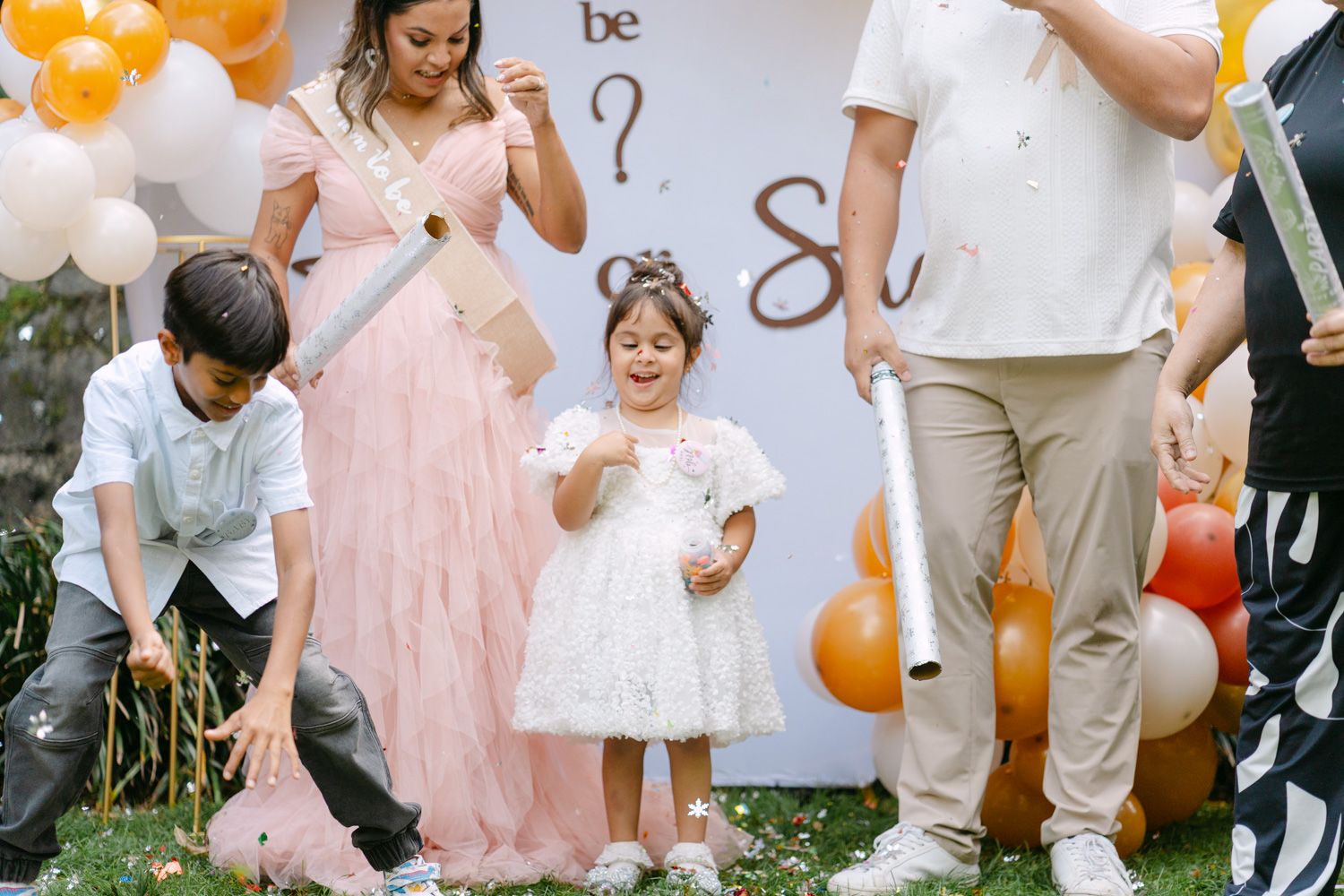 A joyful gender reveal party with children and a woman in a pink gown surrounded by gold and white balloons outdoors.