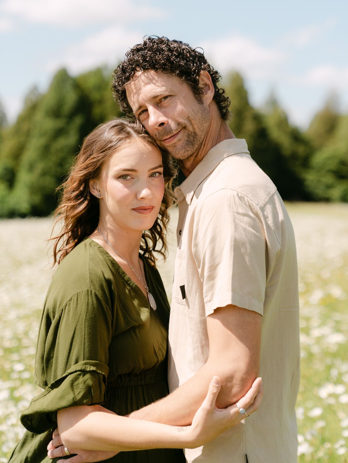 Couple embracing in a sunny wildflower field, woman in green dress, man in beige shirt, with evergreen trees behind.