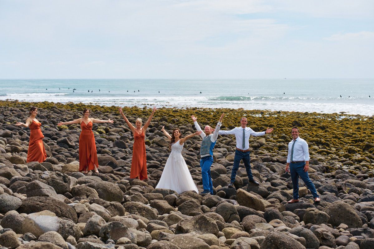 Wedding party celebrating on rocky beach, bride in white, bridesmaids in coral dresses, groomsmen in blue, arms raised joyfully.