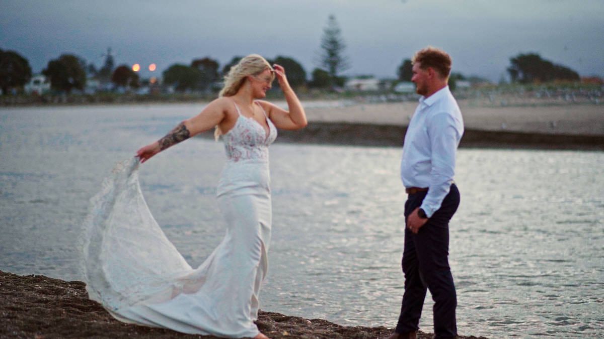 Bride in white gown and groom in dark suit pose romantically on rocky shoreline at dusk near waterfront.