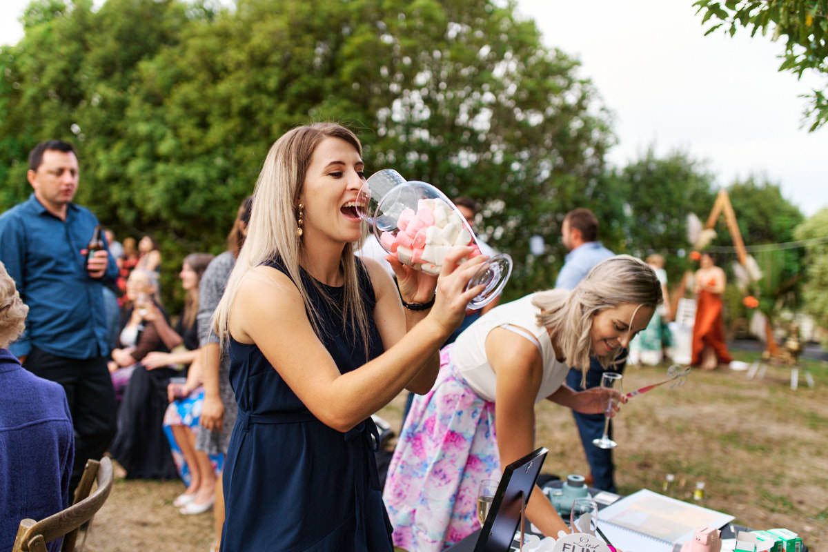 Woman drinking from large glass at outdoor wedding reception with guests socializing in background.