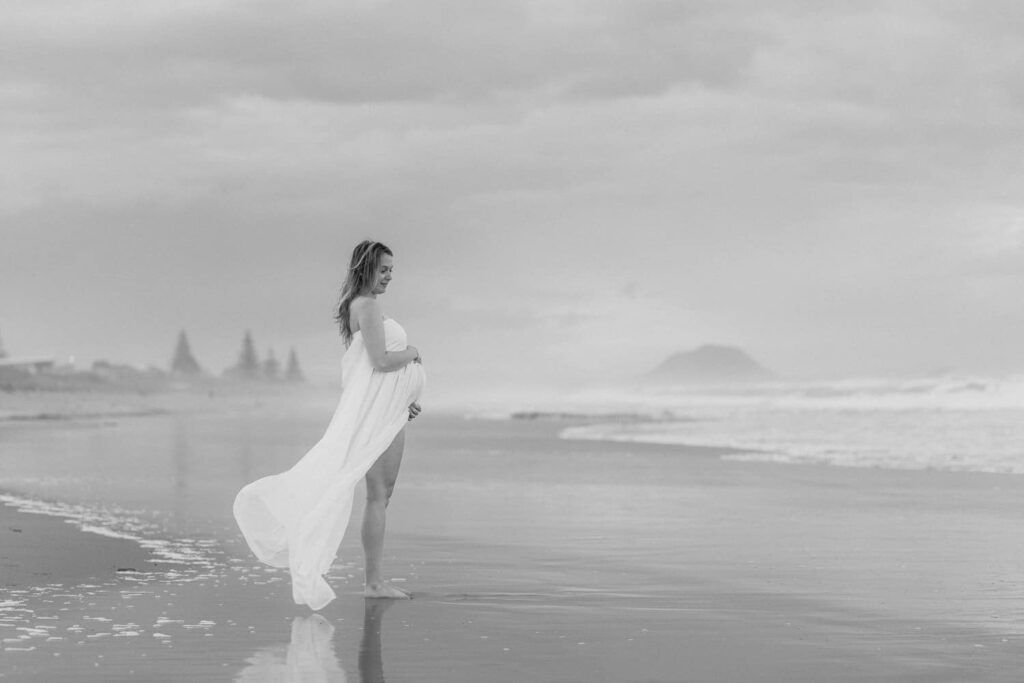 Pregnant woman in flowing white dress standing at ocean's edge on a beach in a black and white photo.