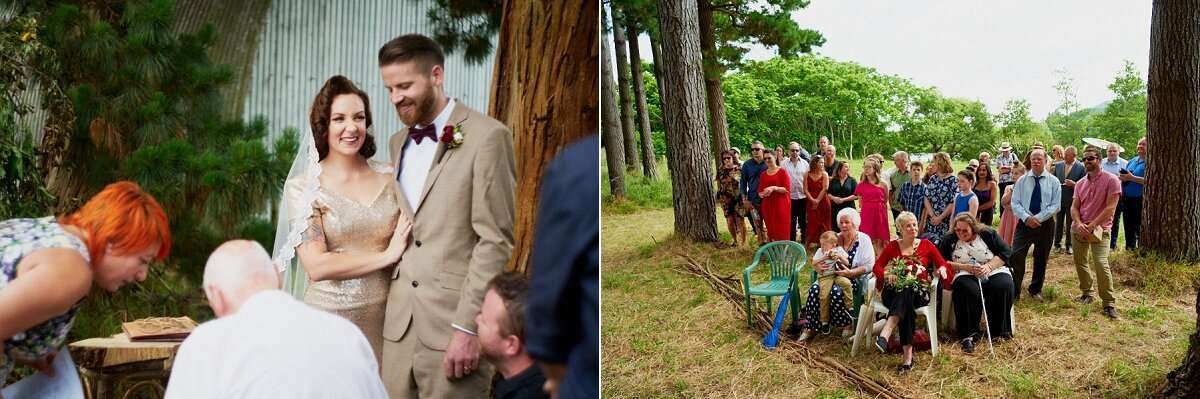 Couple smiles during outdoor wedding ceremony beside tall trees with guests gathered around.