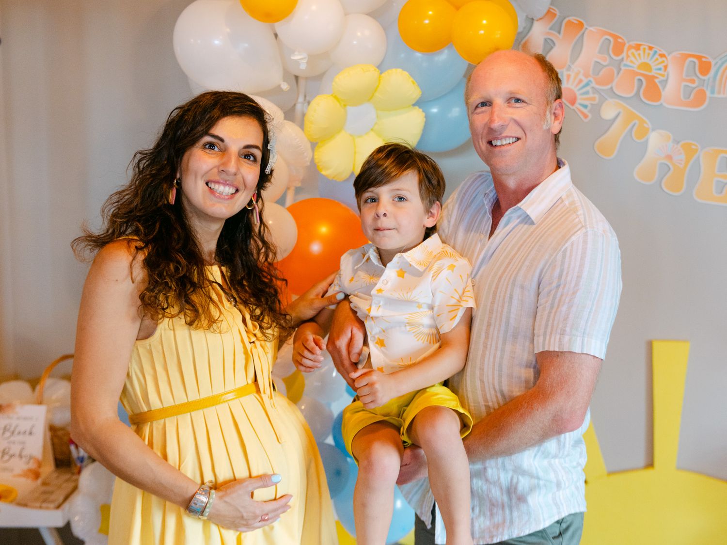 Pregnant woman in yellow dress with husband and young son at a sunny baby shower with balloons.