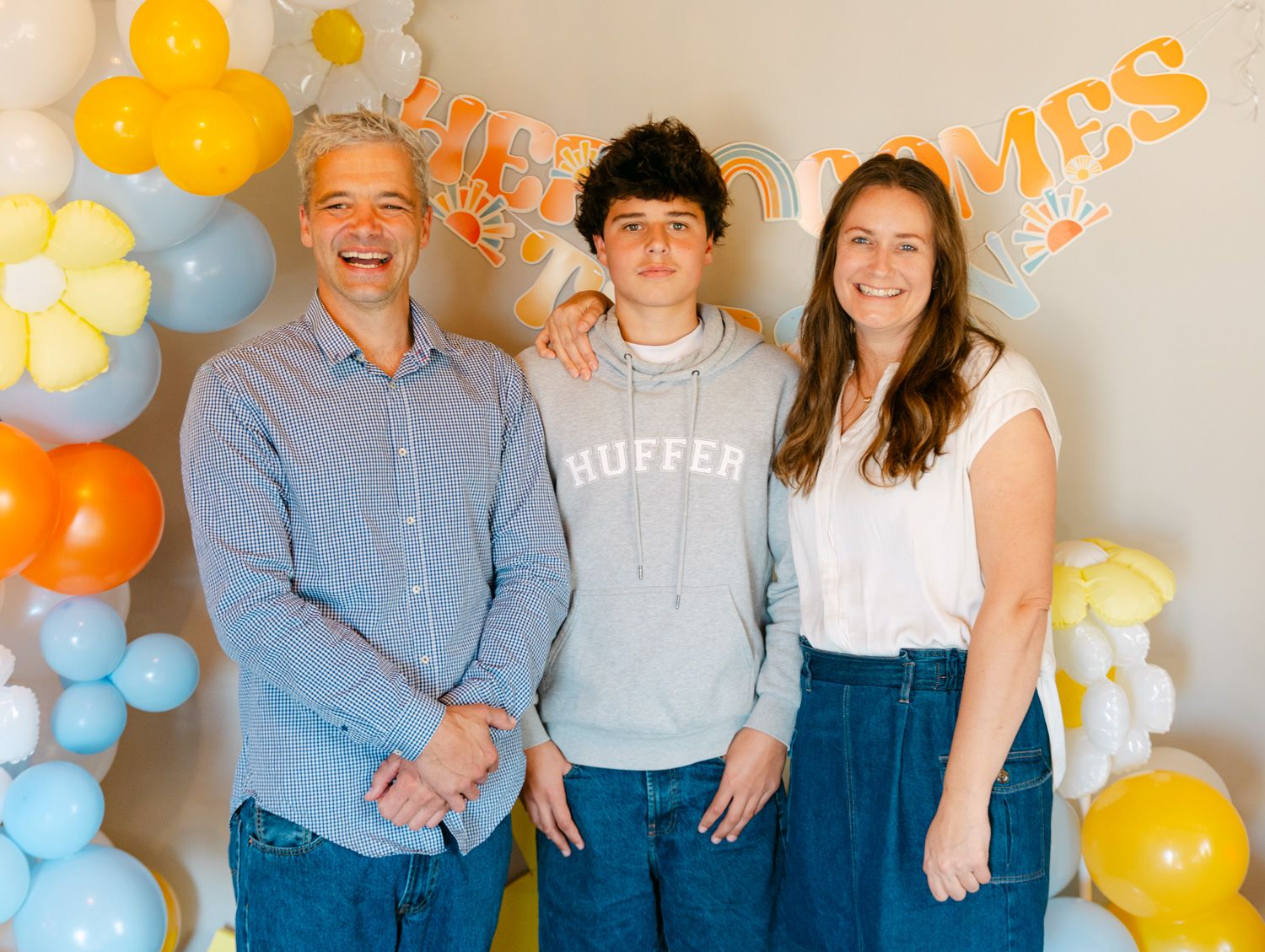 Family of three smiling together at a celebration with colorful balloon decorations and a festive banner backdrop.