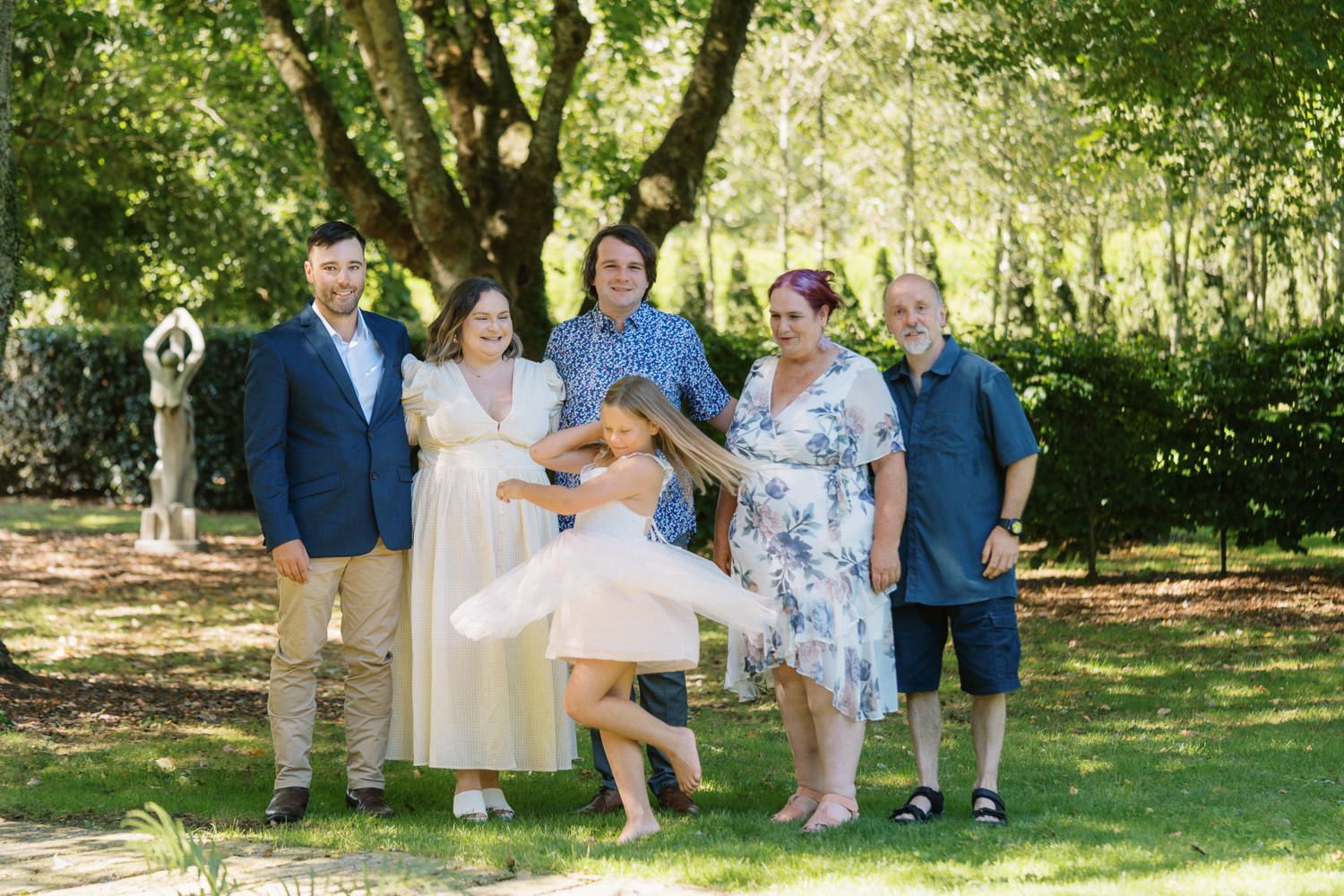 Family group photo outdoors on green lawn with trees, adults and children smiling, one child twirling in white dress.