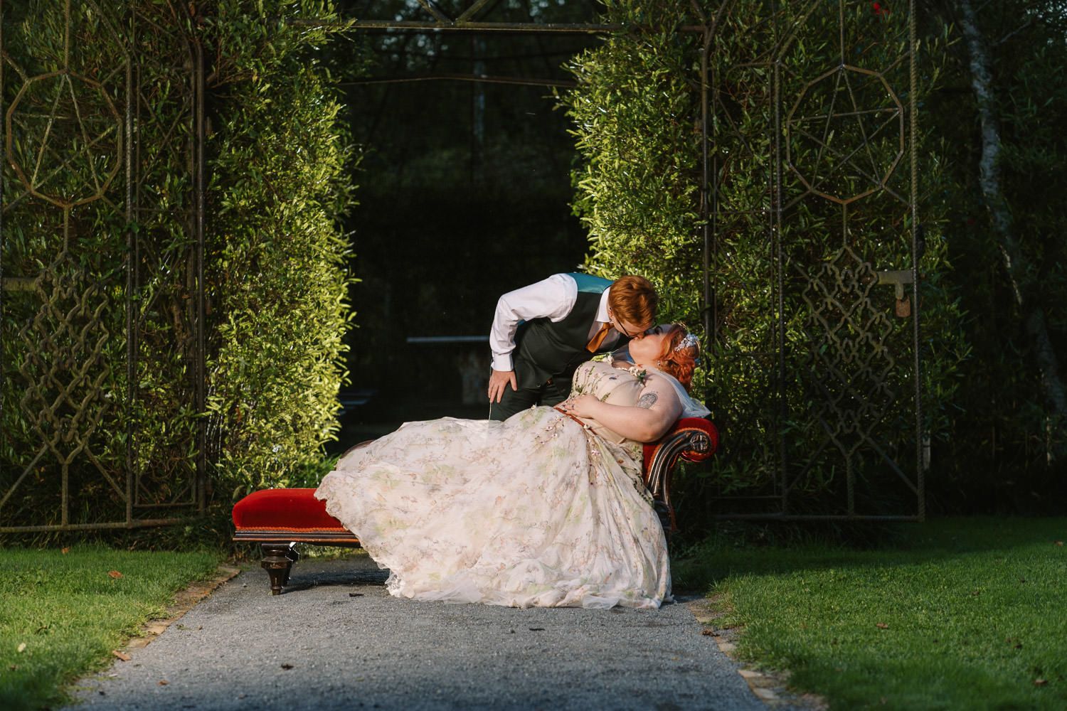 Bride and groom share a romantic dip kiss on a red bench surrounded by lush green hedges in a garden.