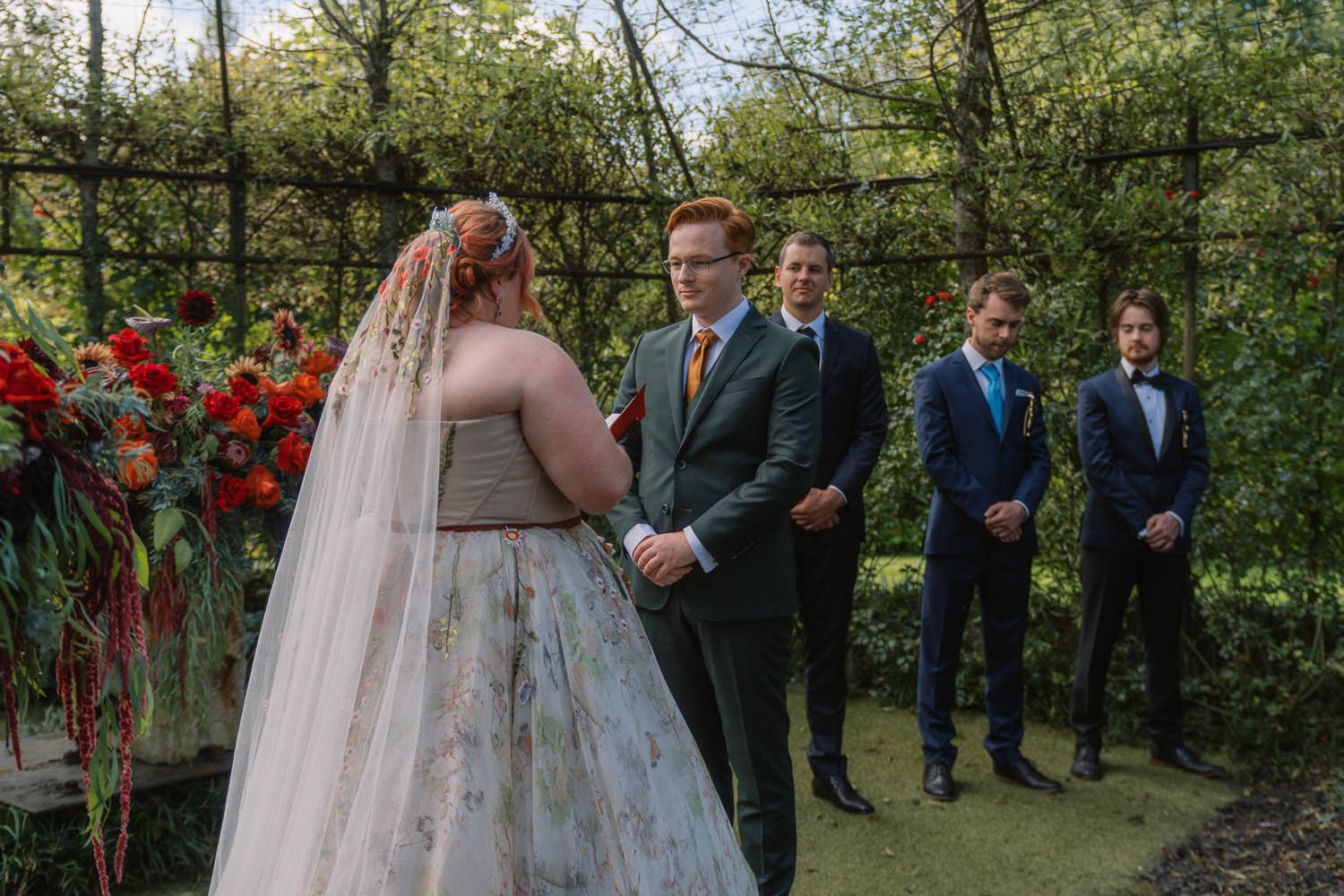 A bride in a colorful gown and veil exchanges vows with a groom in a green suit at an outdoor garden wedding.