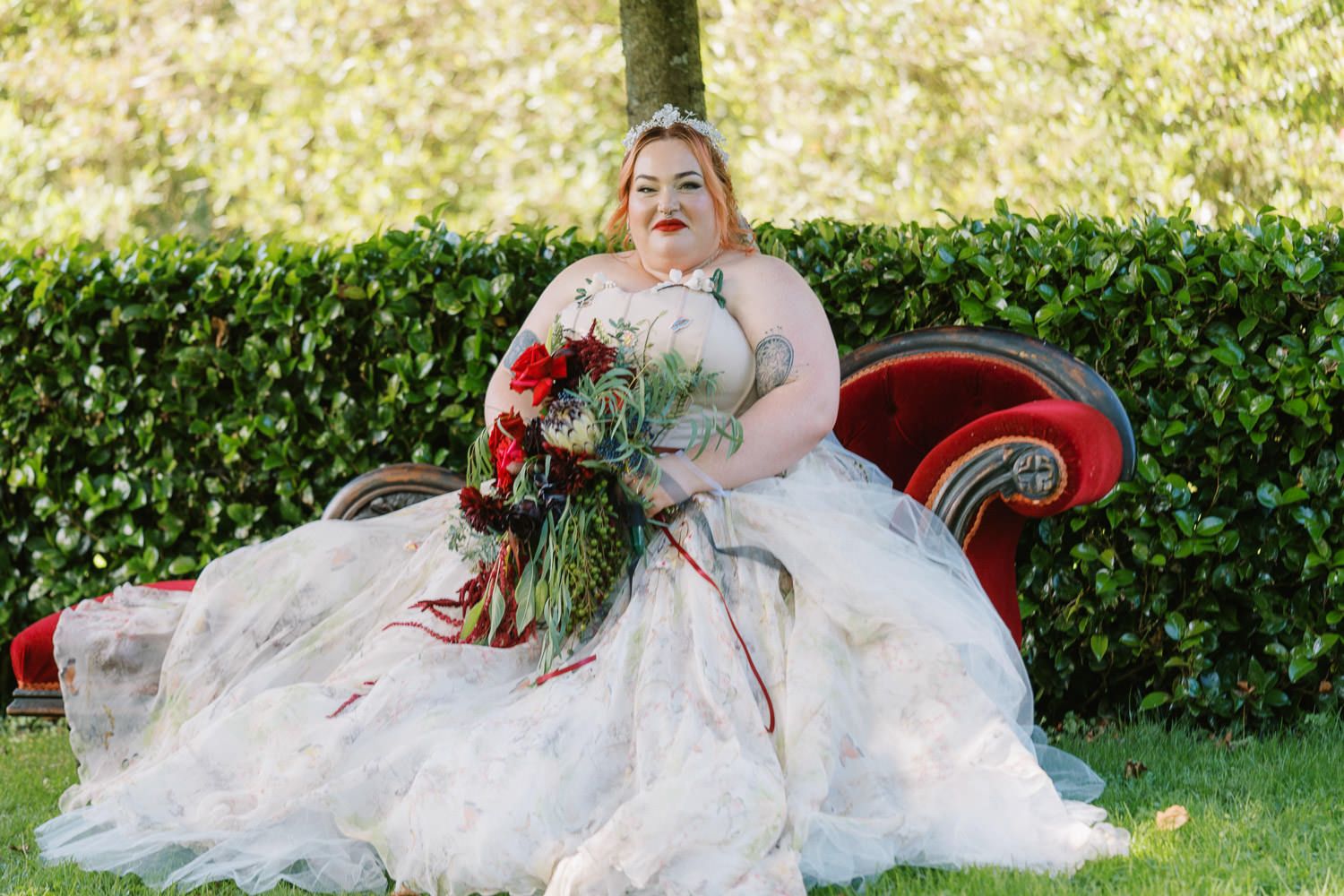 Plus-size bride in white ball gown holding red bouquet, seated on red velvet sofa outdoors with green hedge backdrop.