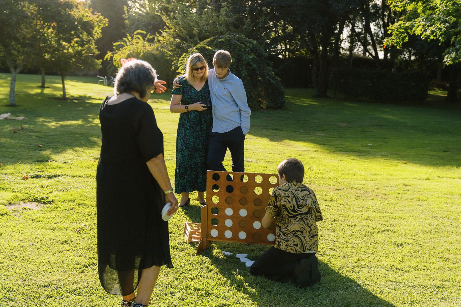 Group of people playing giant Connect Four game on green lawn at outdoor garden party in warm sunlight.