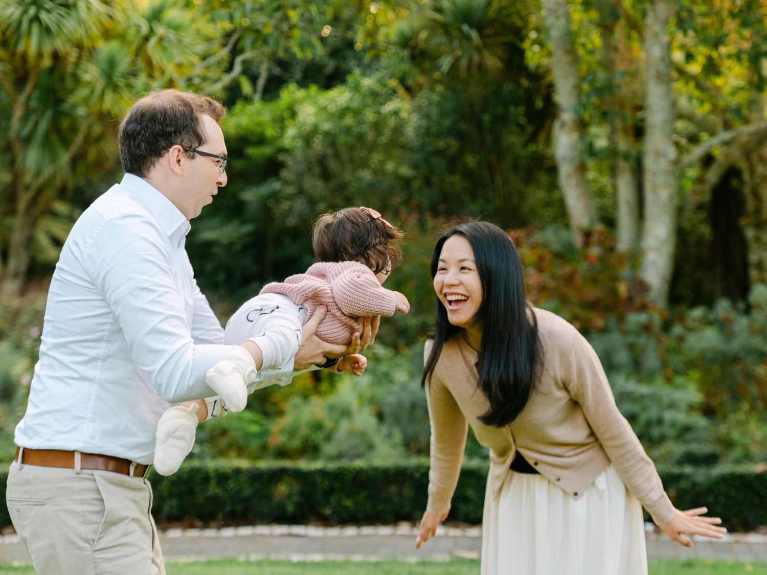 Happy family playing outdoors in a lush green park, father lifting baby while mother laughs joyfully.