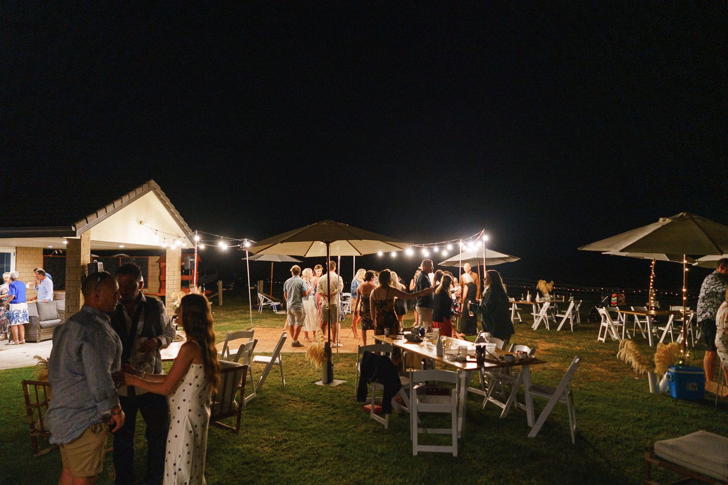 Outdoor evening wedding reception with string lights, white chairs, and guests mingling on a lawn under a night sky.