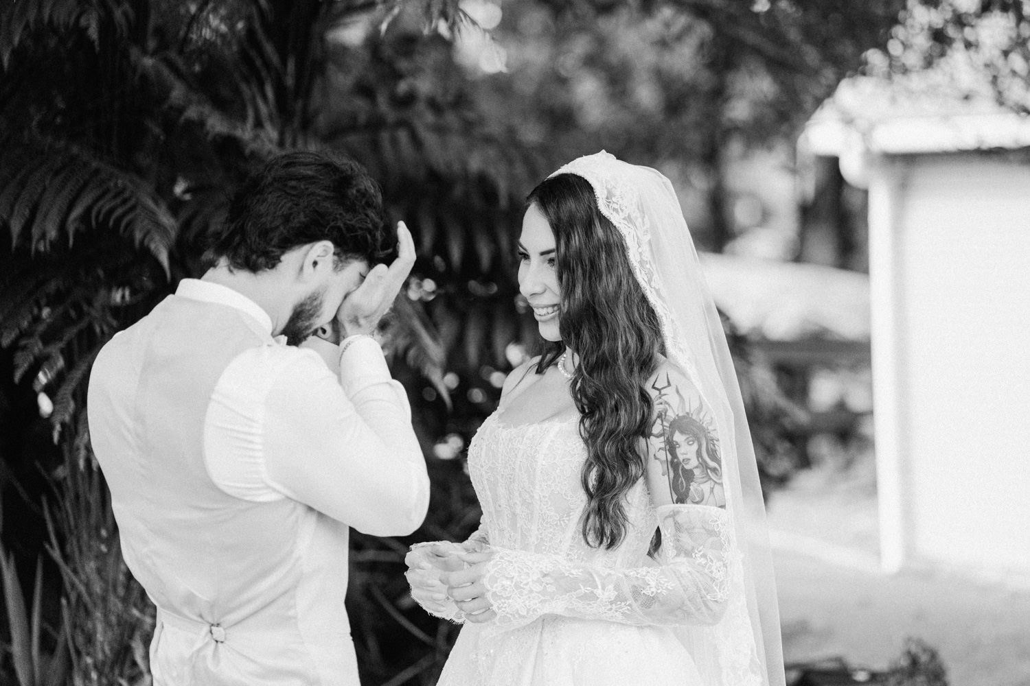 Emotional black and white wedding photo of groom wiping tears while bride smiles tenderly.
