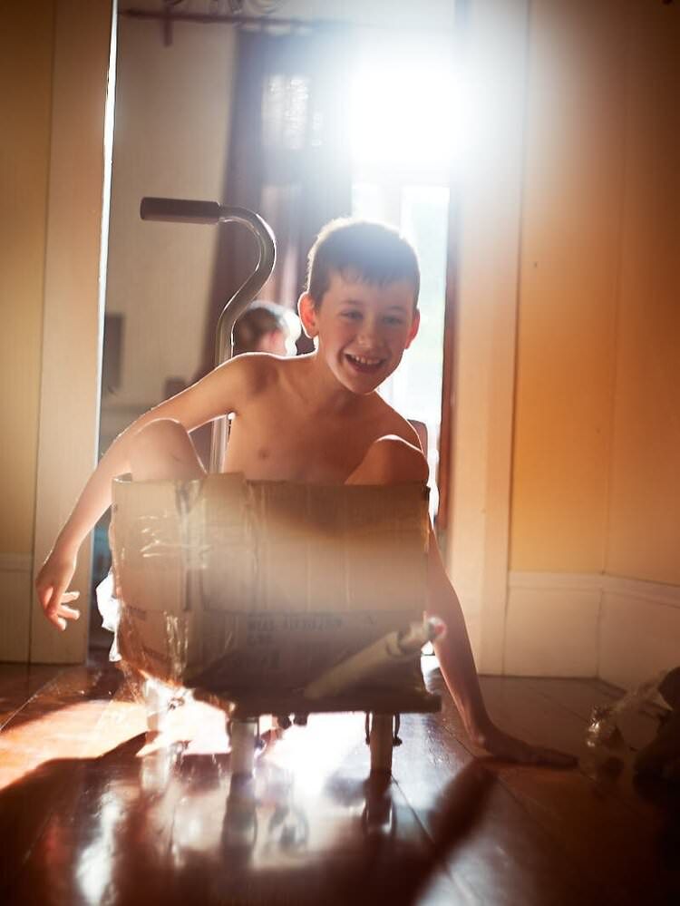 A smiling shirtless boy plays with a wooden barrel on a shiny hardwood floor in a bright room.