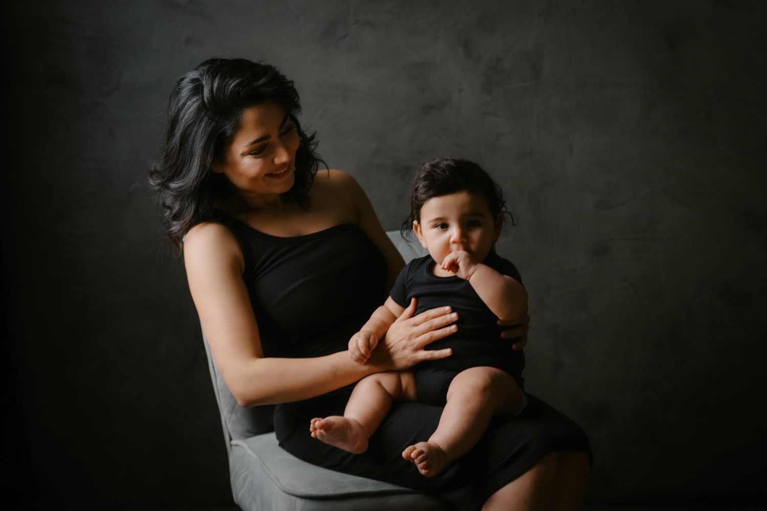 A mother in a black dress holds her baby in a dark studio portrait setting.