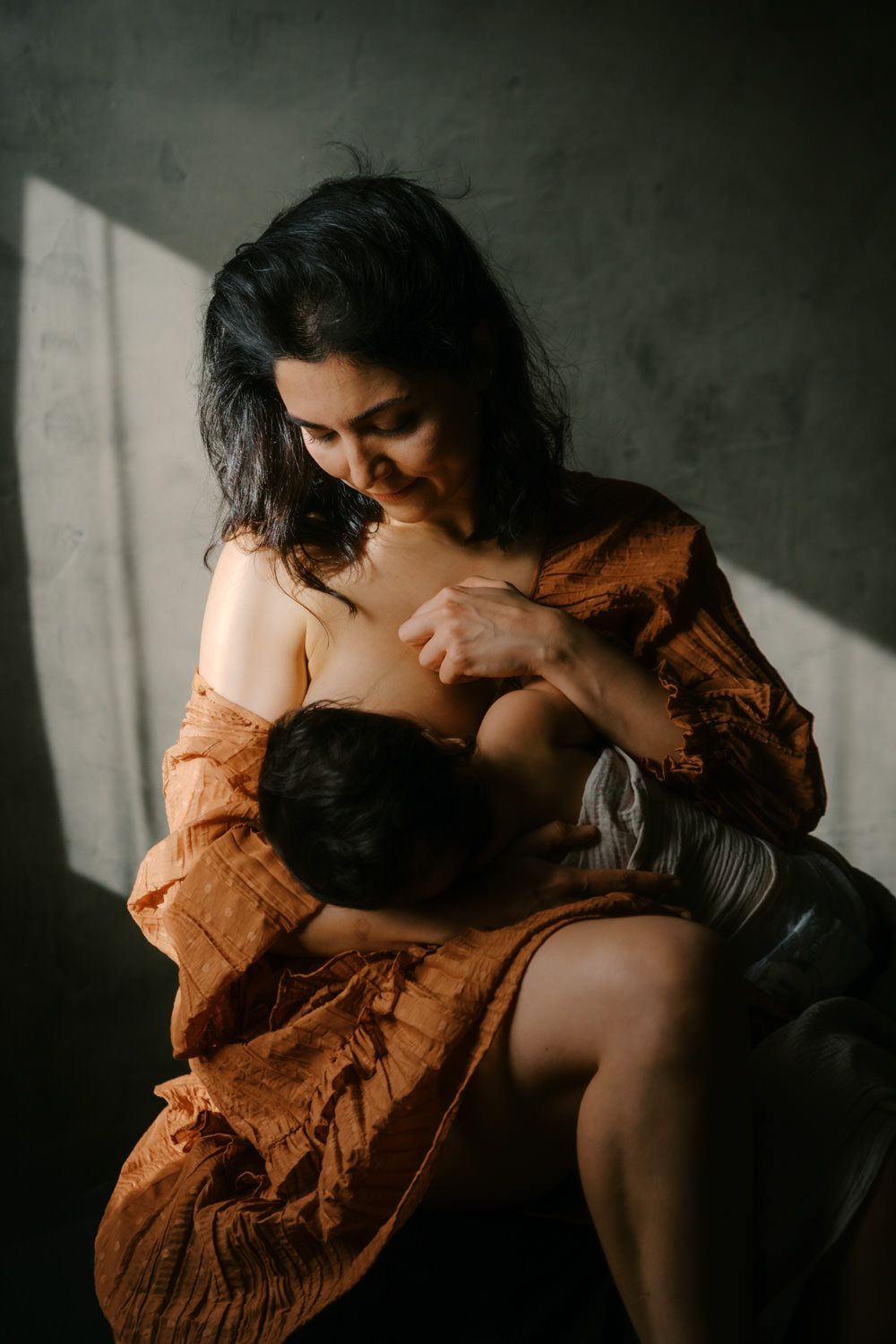 Mother breastfeeding her baby in warm natural light, wearing an earthy orange blouse in a moody studio setting.
