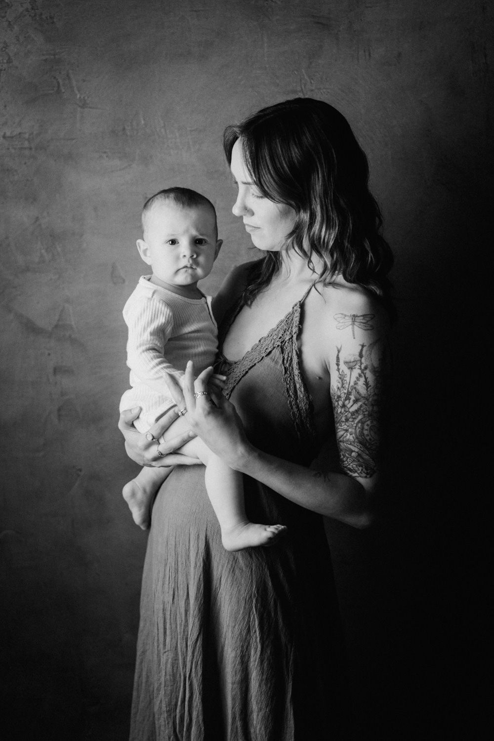 Black and white portrait of a mother with tattoos holding her baby, gazing down lovingly in a studio setting.