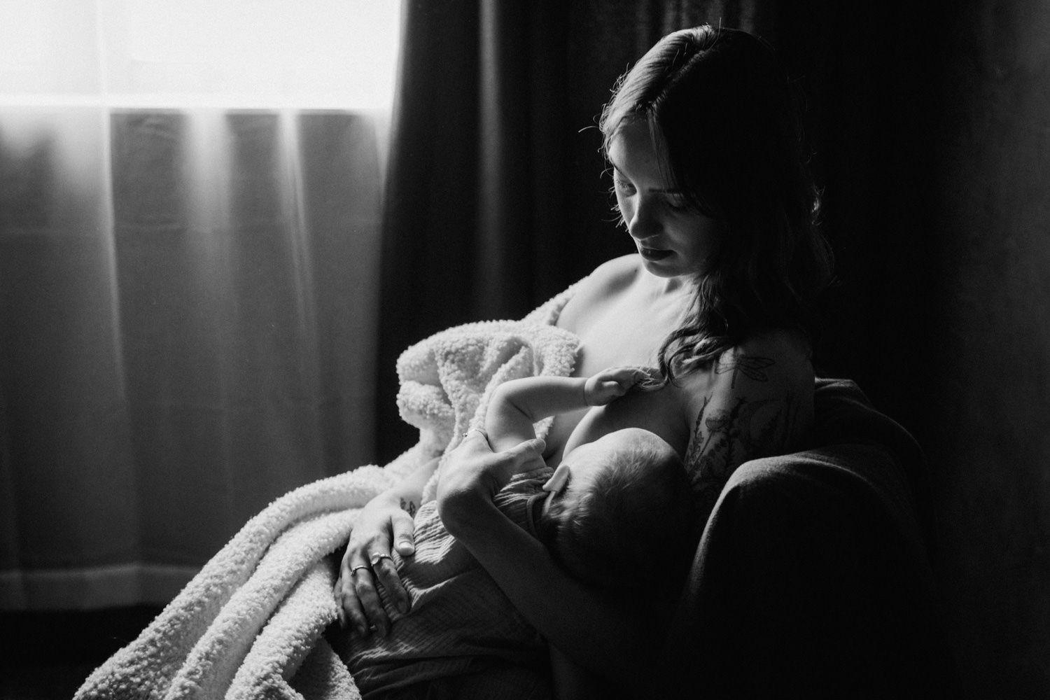 Black and white photo of a mother tenderly cradling her newborn baby while seated by a curtained window.