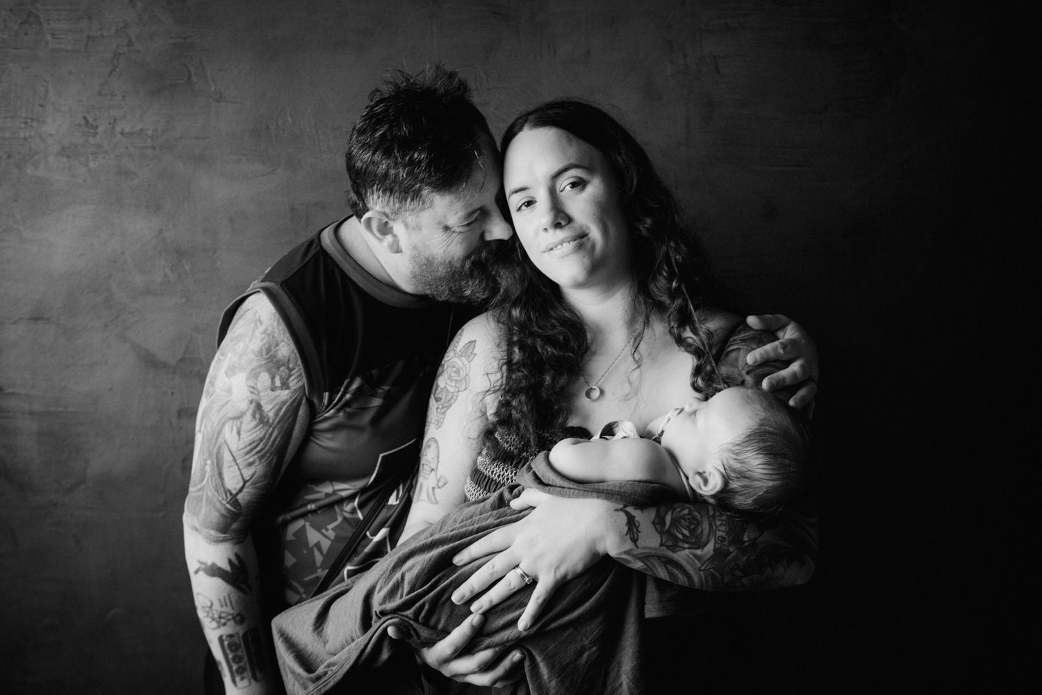 Black and white family portrait of tattooed parents tenderly holding their newborn baby in a studio setting.
