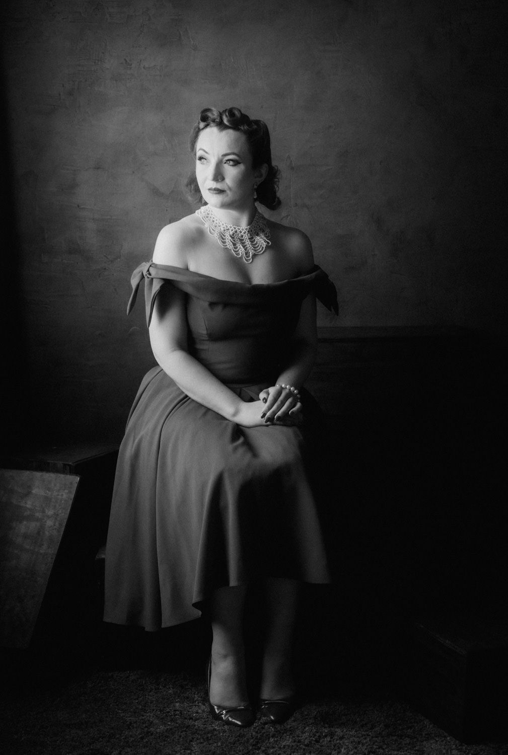 Black and white portrait of a woman in an off-shoulder dress with pearl necklace, seated elegantly in dramatic studio lighting.