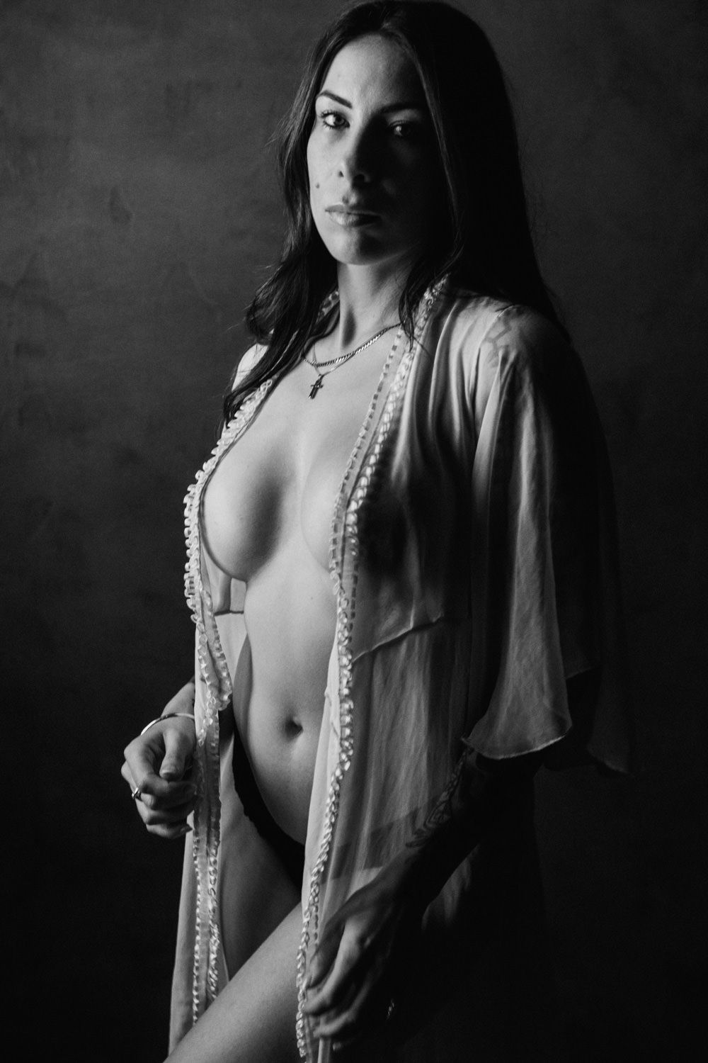 Black and white boudoir portrait of a woman in sheer open robe posing confidently against dark background.