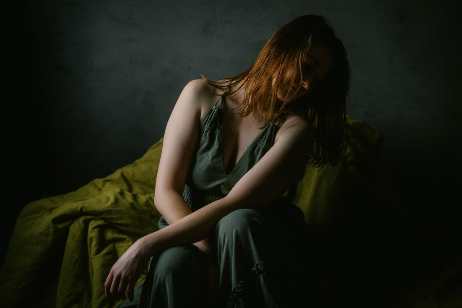 A young redhead woman in a green dress sits pensively with head bowed in moody, dramatic studio lighting.
