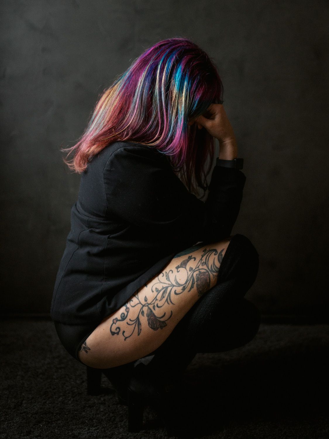 Person with colorful rainbow hair crouching in dark studio, showing detailed floral tattoo on leg, wearing black outfit.