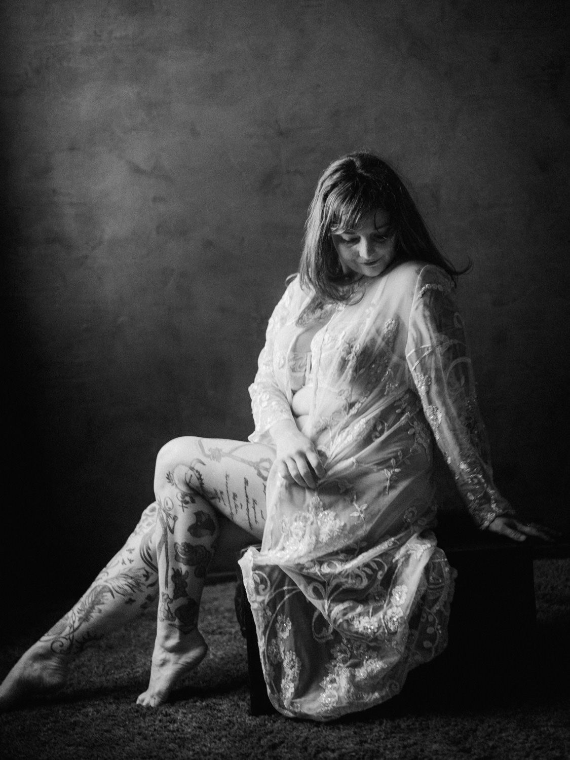 Black and white maternity photo of a pregnant woman in lace dress sitting gracefully in a studio setting.