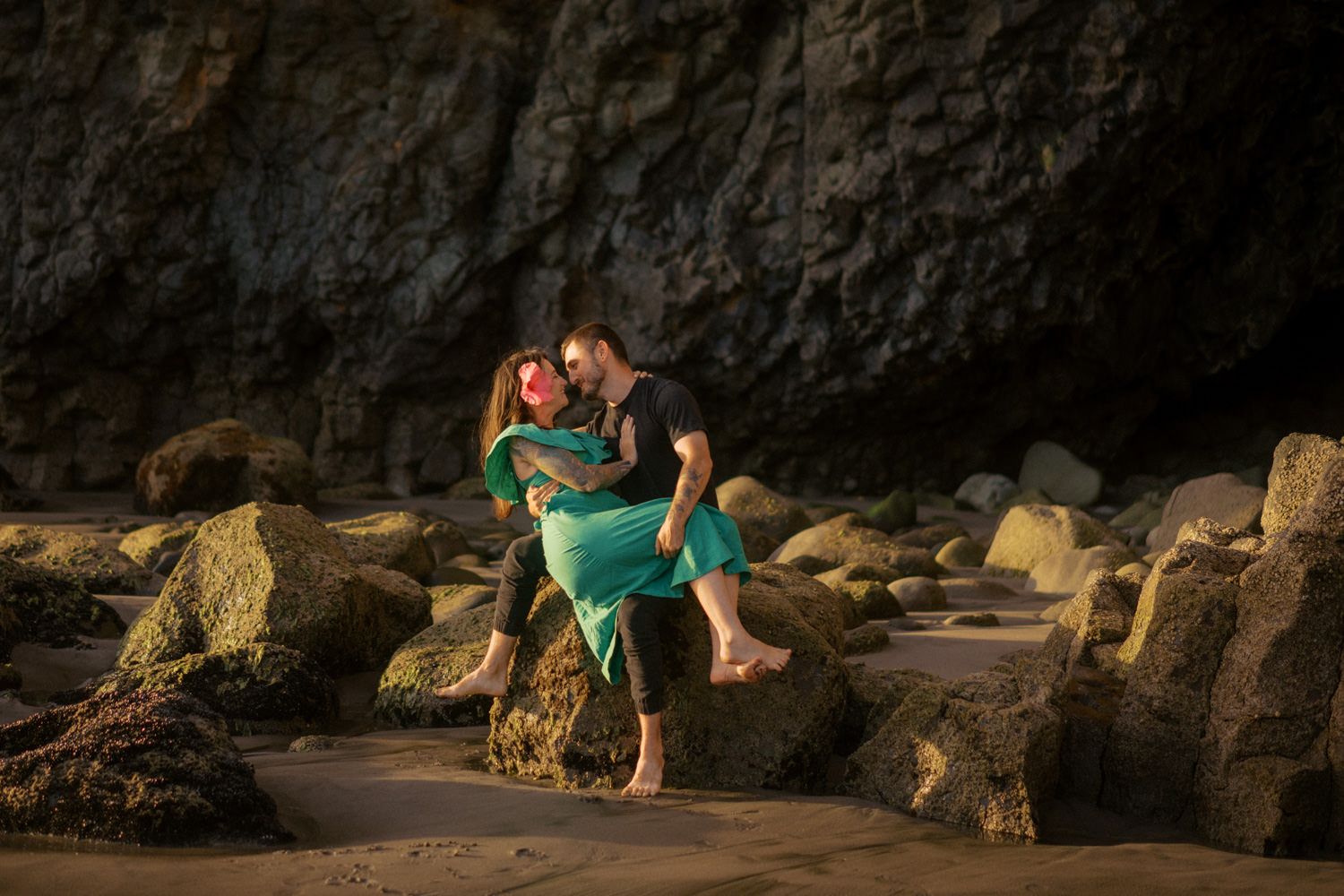 Romantic couple embracing on rocky beach near dramatic cliffs at golden hour, woman in teal dress.