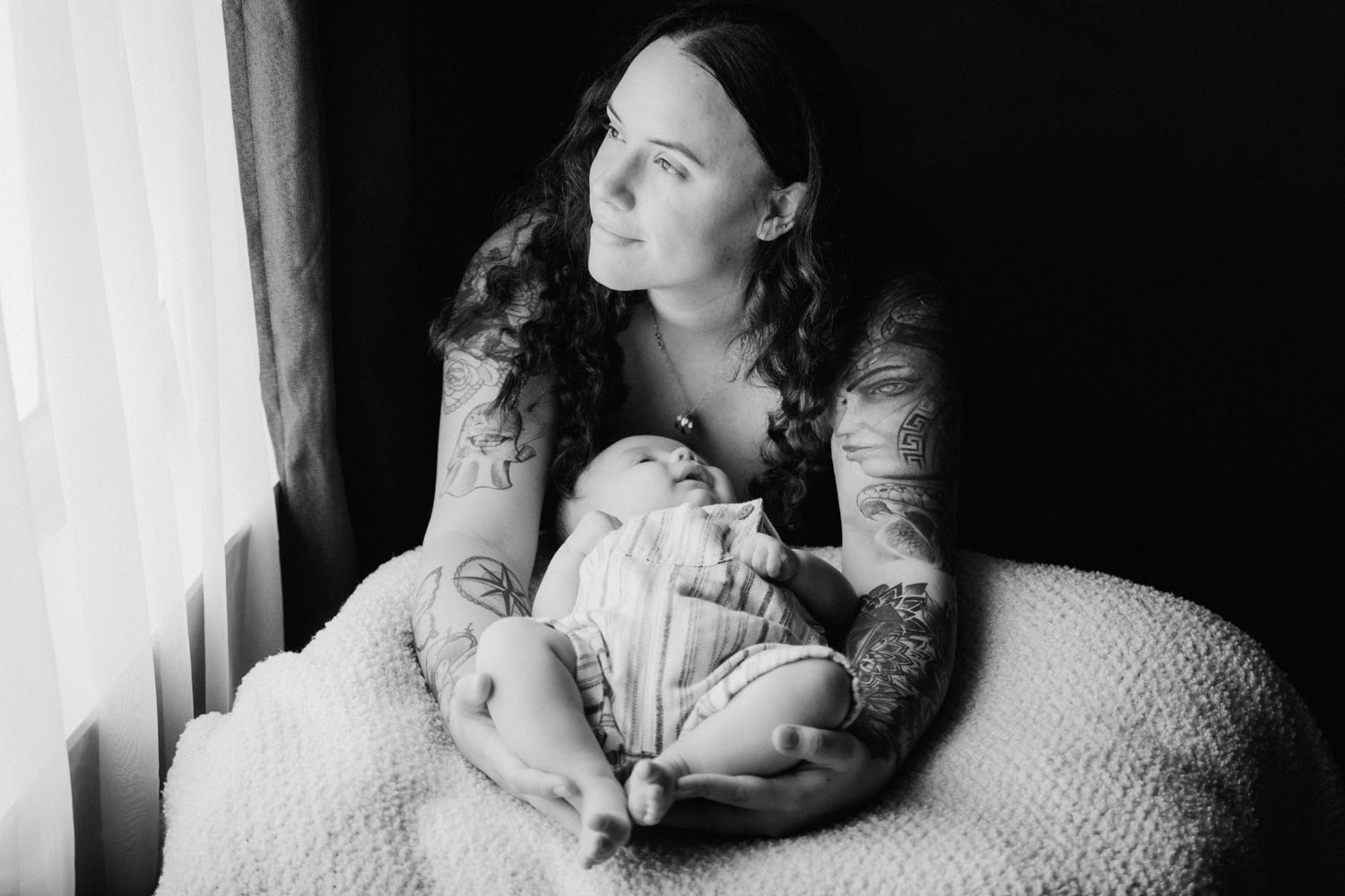 Black and white photo of tattooed mother cradling sleeping newborn baby by window light.