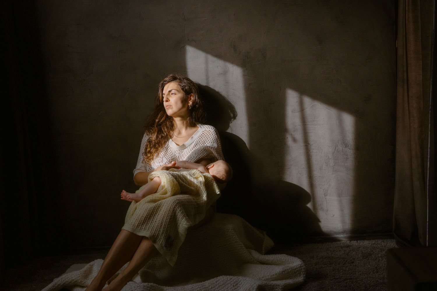 A mother sits on the floor in soft window light, cradling her newborn baby in a serene, warmly lit room.