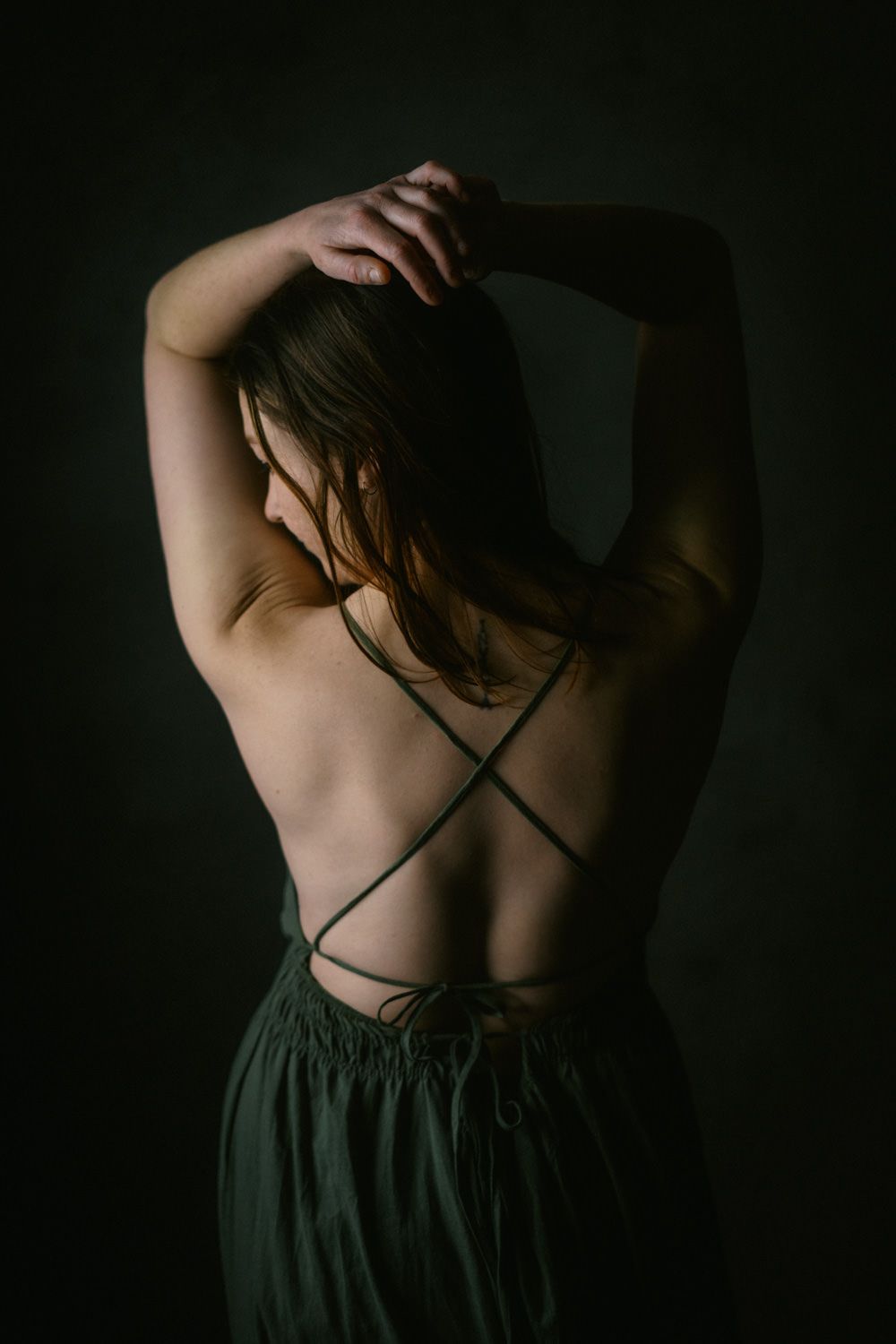 Woman in backless green dress with crossed straps poses with arms raised against dark background.