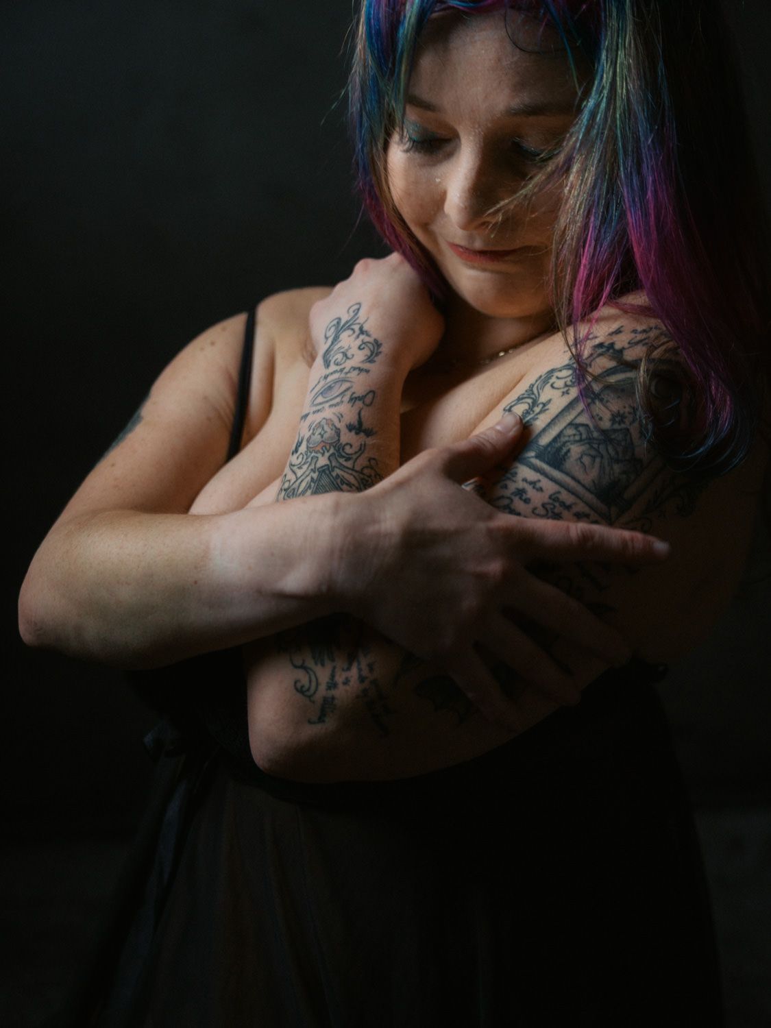 Woman with colorful blue and pink hair and tattoos on her arms crosses them over her chest against a dark background.
