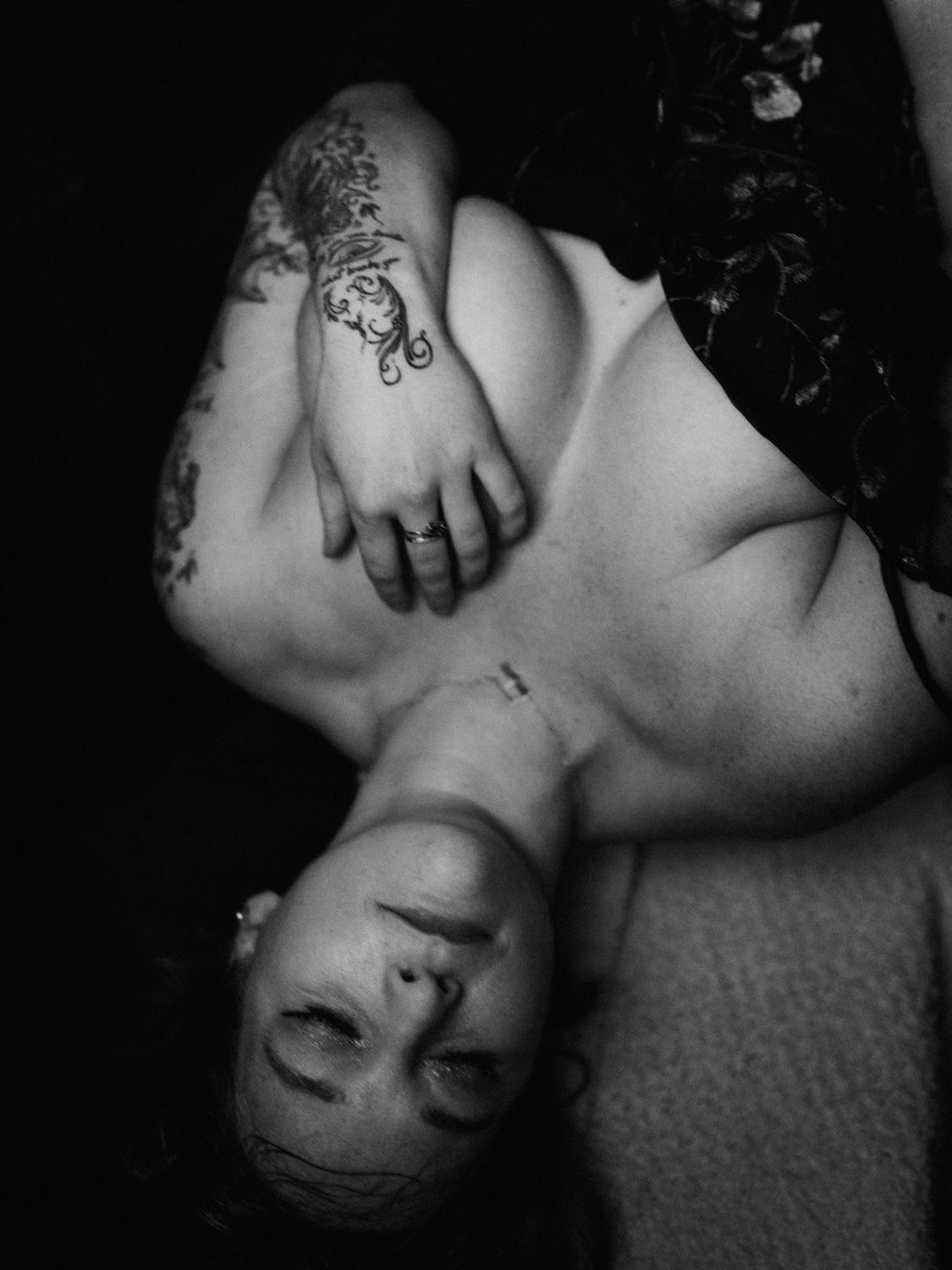 Black and white photo of a woman lying down with tattoos on her arm and hand, wearing dark clothing.