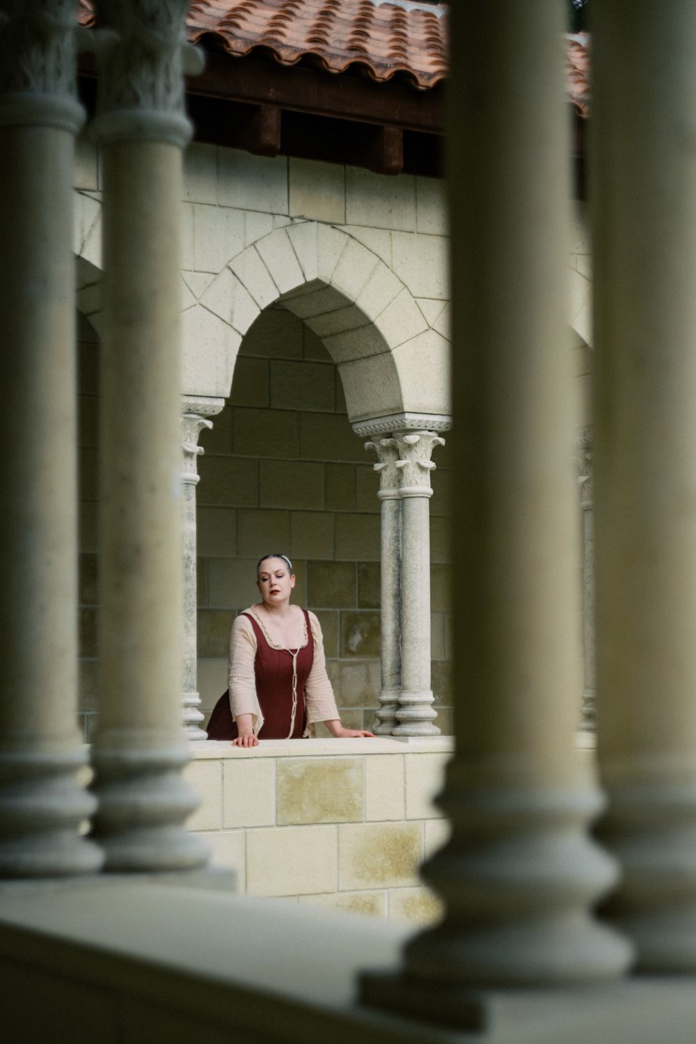 Woman in red dress standing among stone arches and columns of a historic building courtyard.