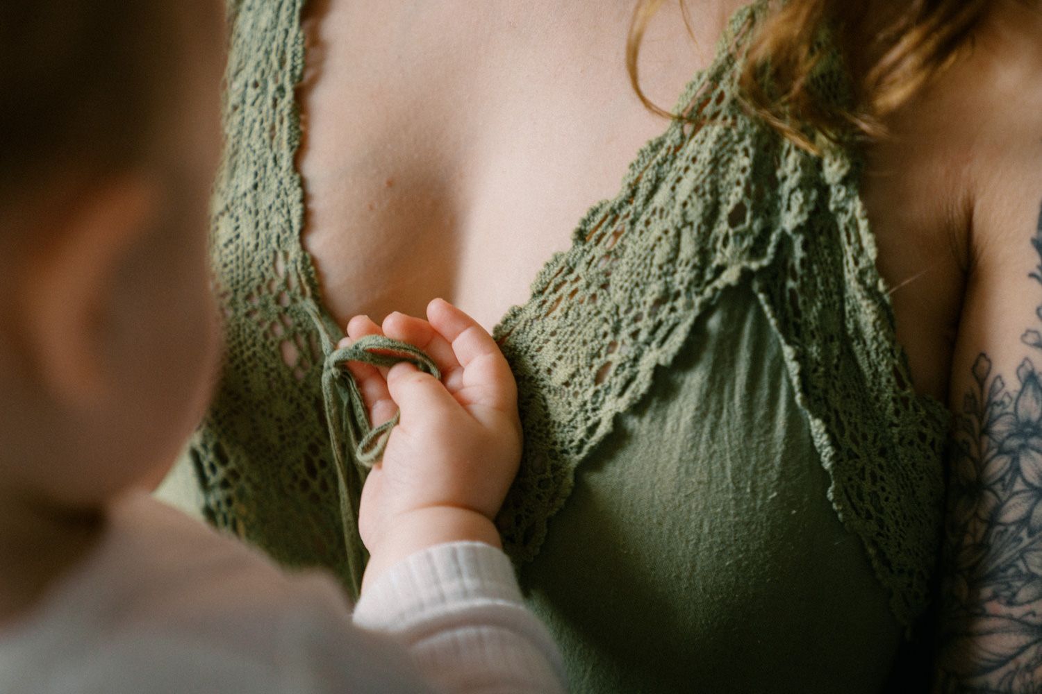 A baby's tiny hand gently touching a woman's green lace bralette, with a tattoo visible on her arm.