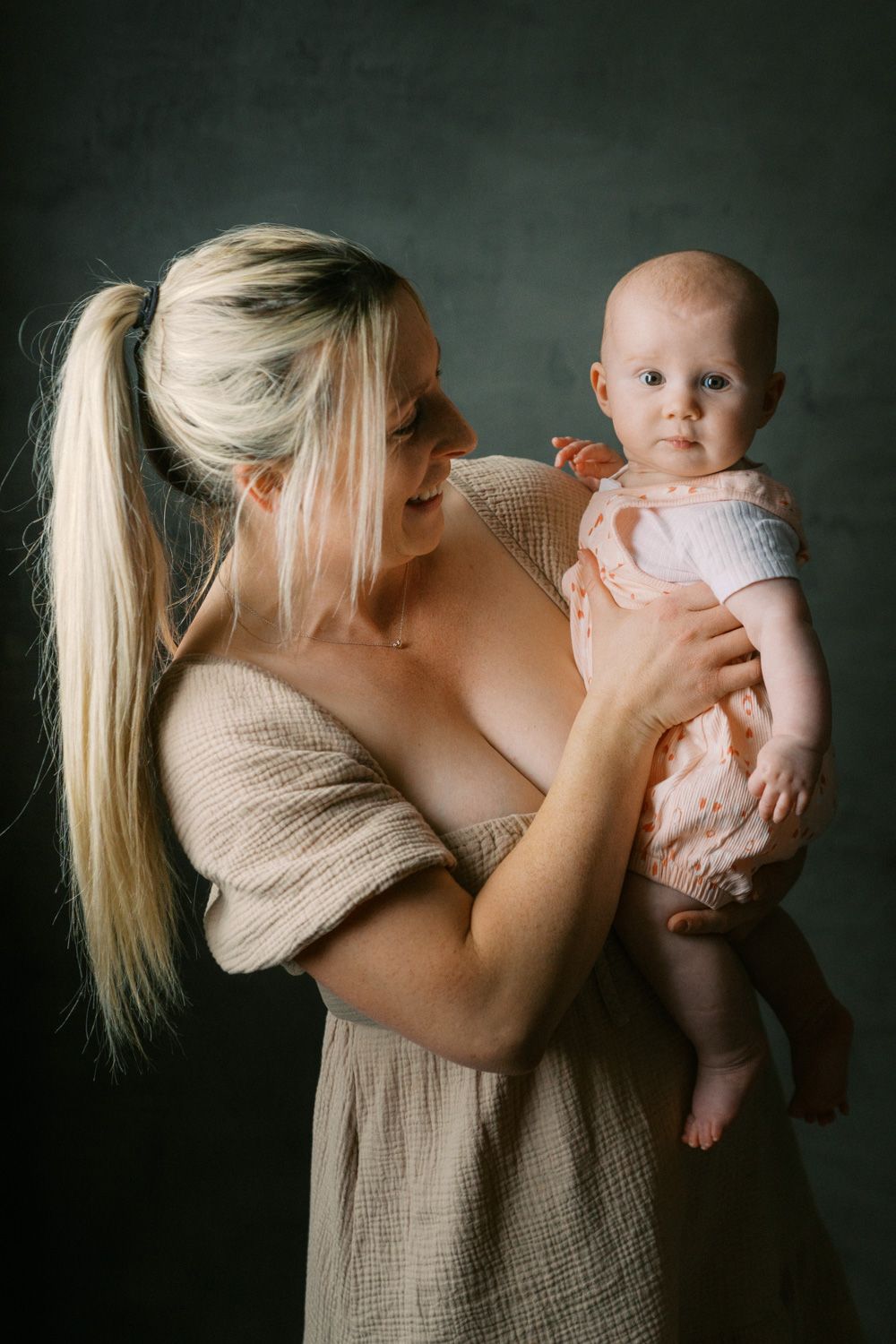 Blonde woman in beige dress lovingly holds a baby in pink outfit against dark background.