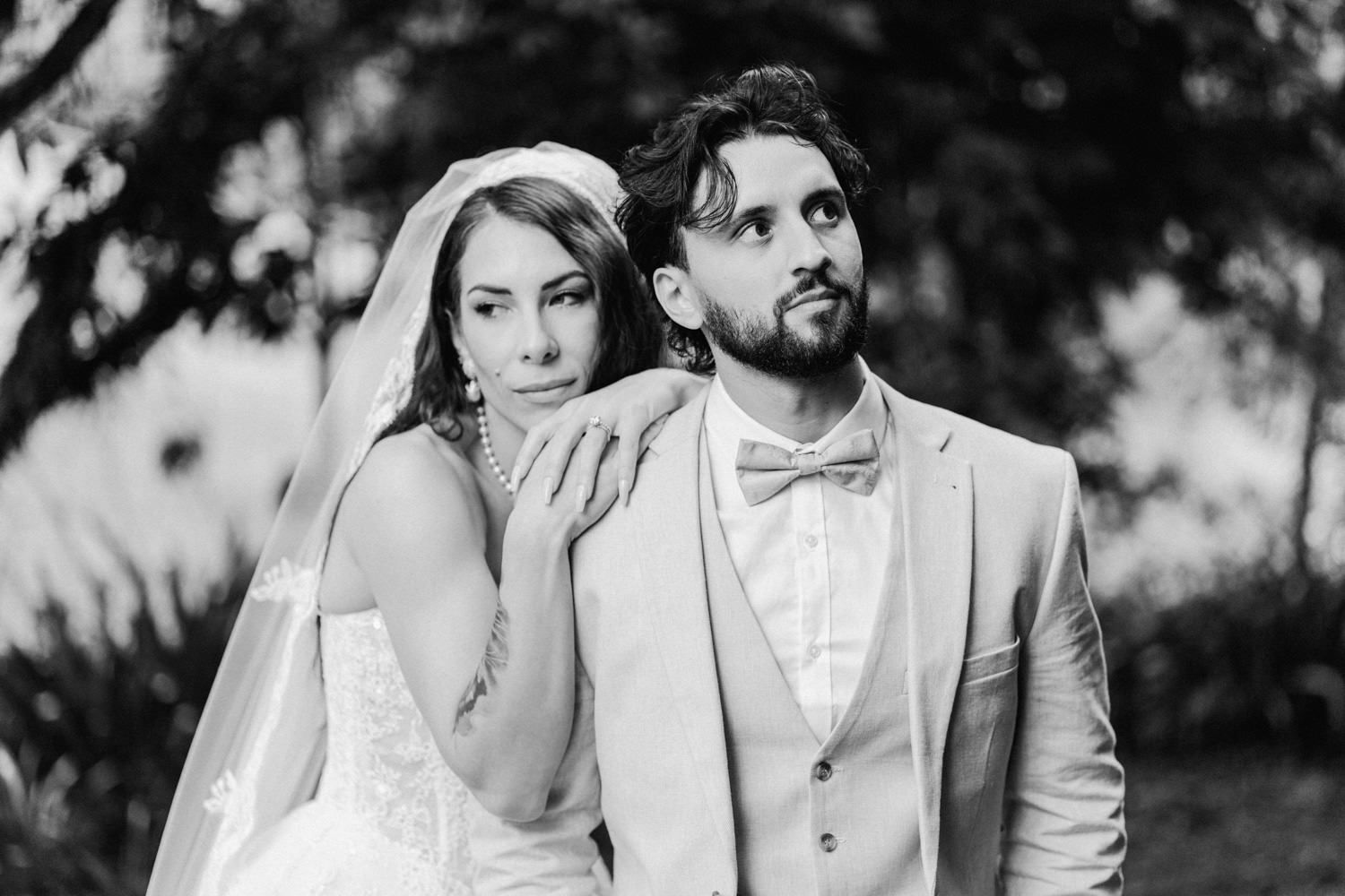 Black and white wedding photo of bride in veil and groom in bow tie suit posing outdoors.