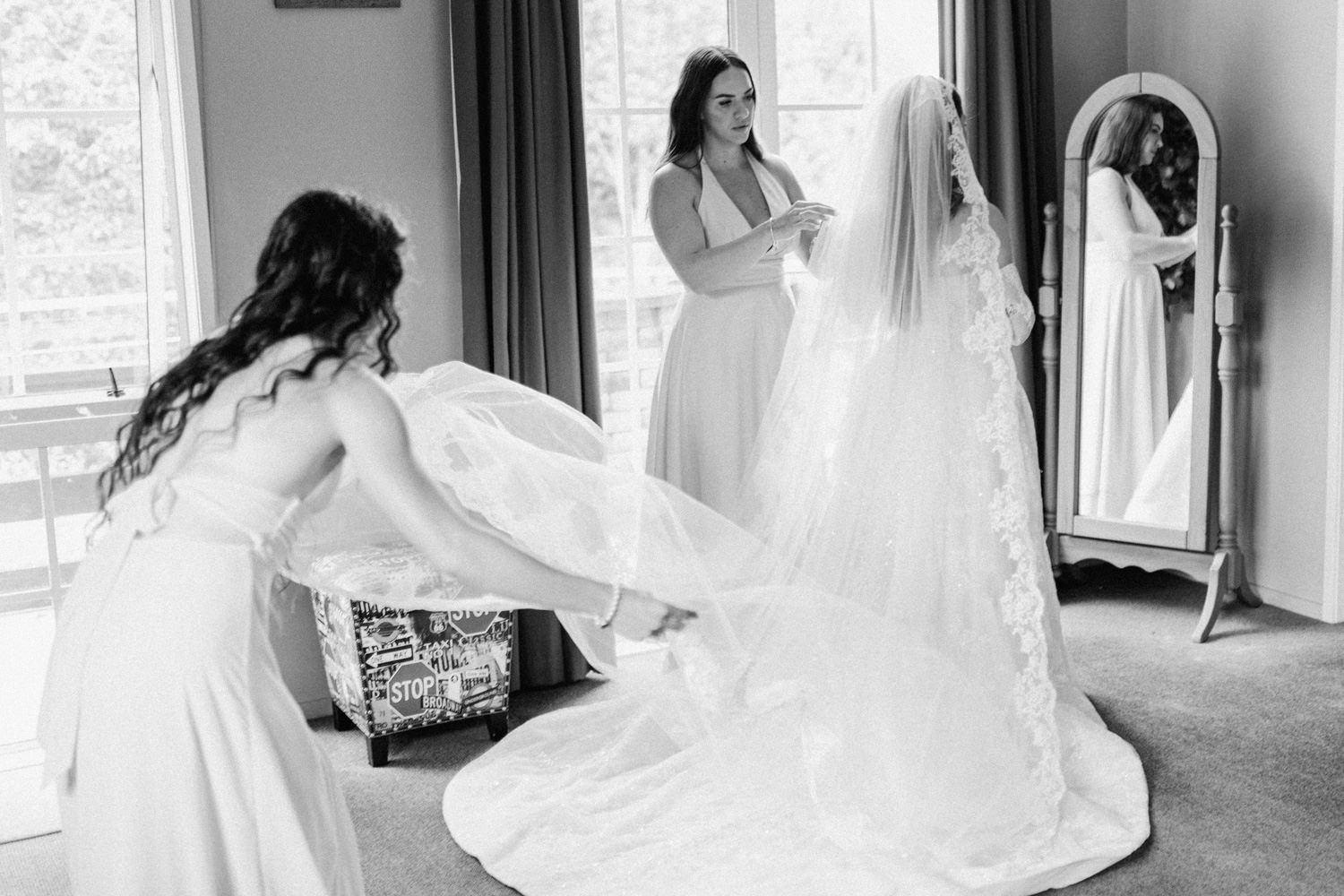Bridesmaids help a bride with her lace wedding gown and veil in a black and white bridal preparation photo.