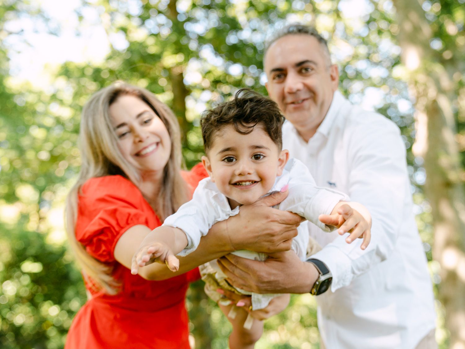 Happy family with parents holding smiling toddler boy outdoors in lush green park setting.