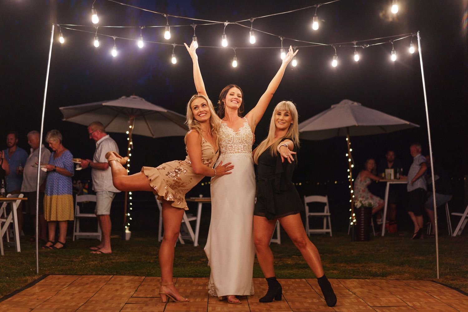 Bride celebrates with two friends under festive string lights at outdoor evening wedding reception.