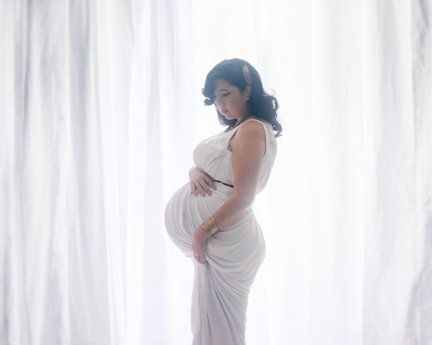 Pregnant woman in white draped gown holding her belly, standing by sheer curtains in a soft maternity portrait.