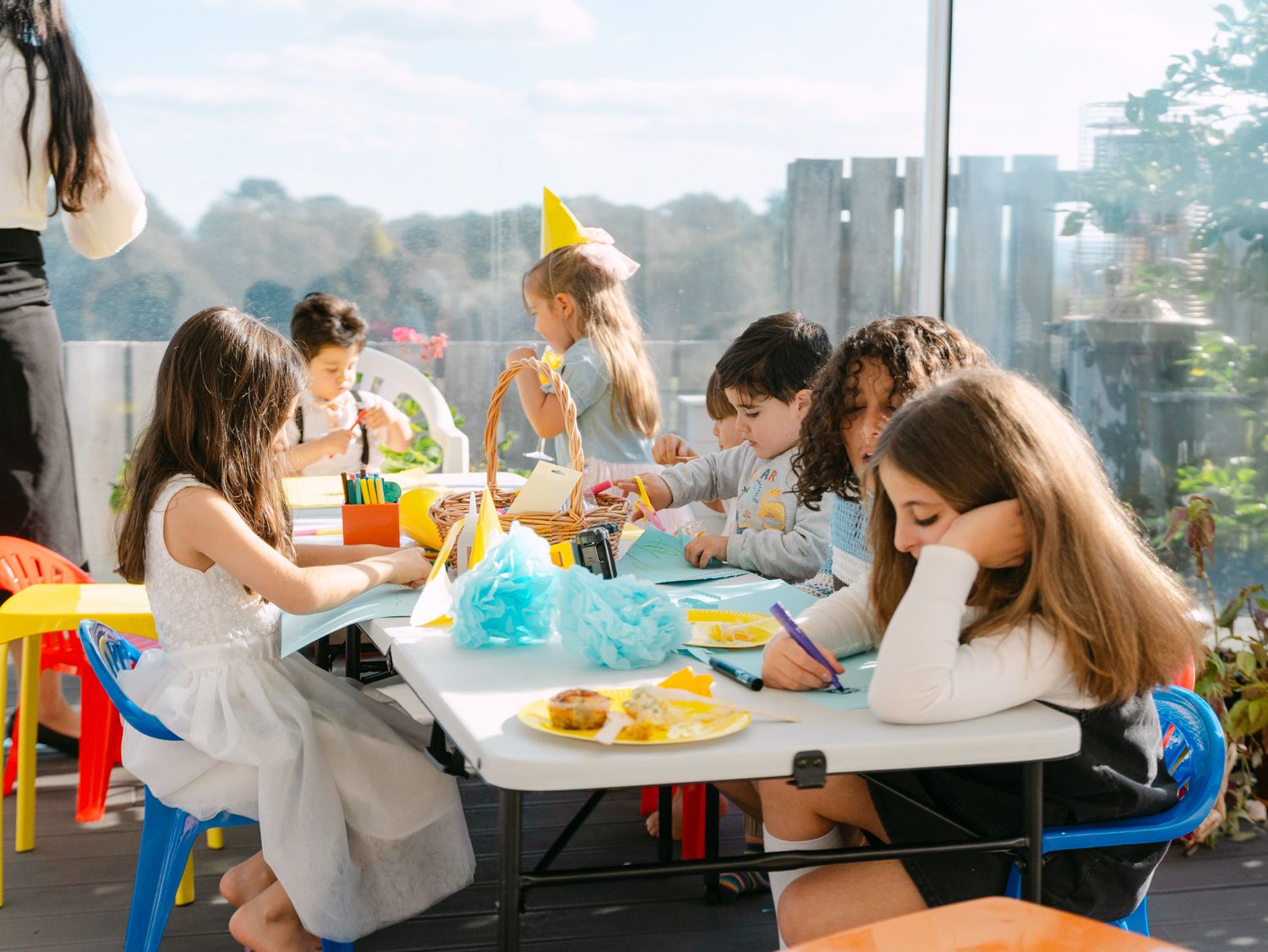 Children enjoying a birthday party craft activity at a white table on a bright indoor patio with colorful chairs.
