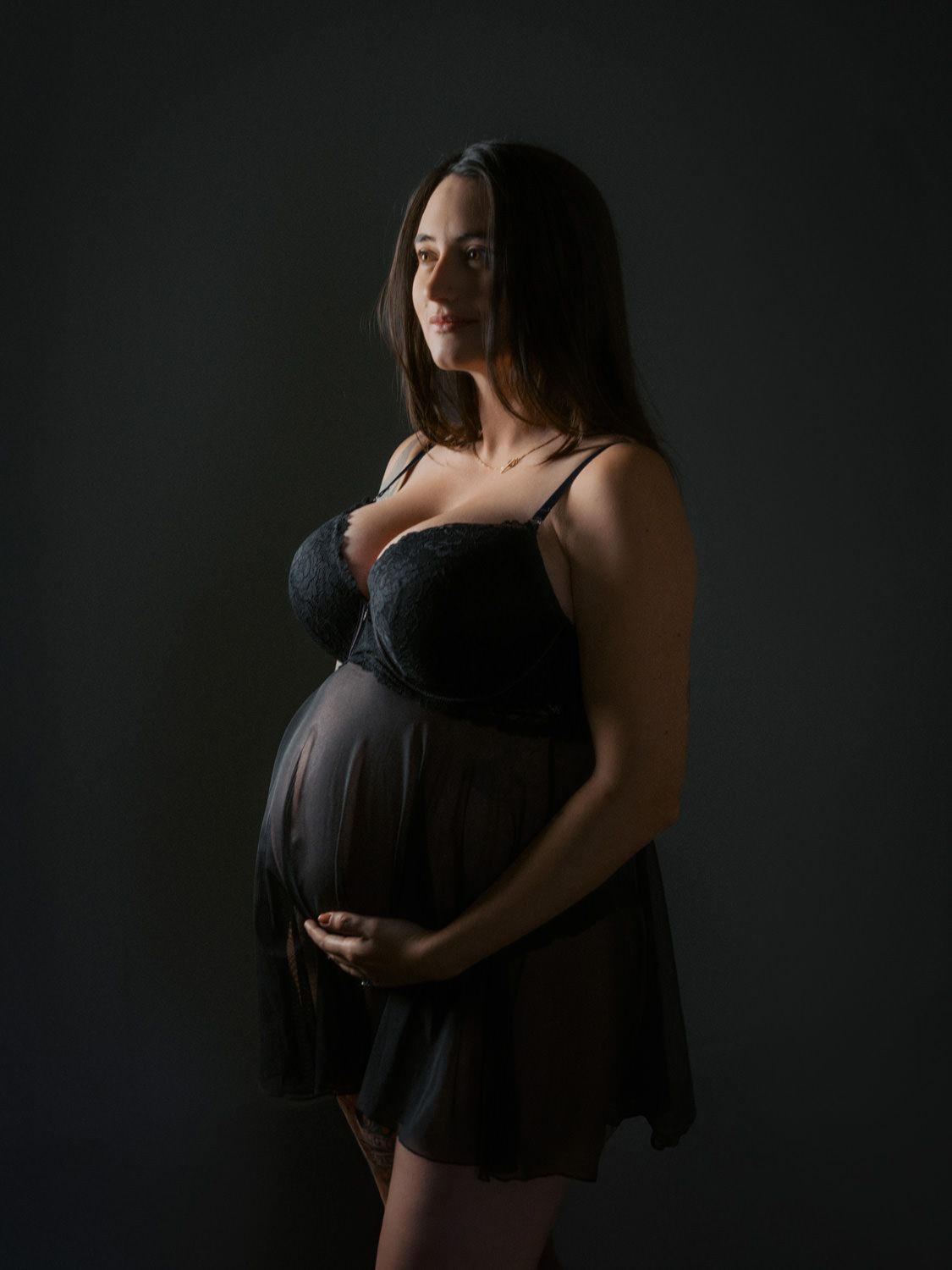 Pregnant woman in black lingerie cradling baby bump against dark studio background.