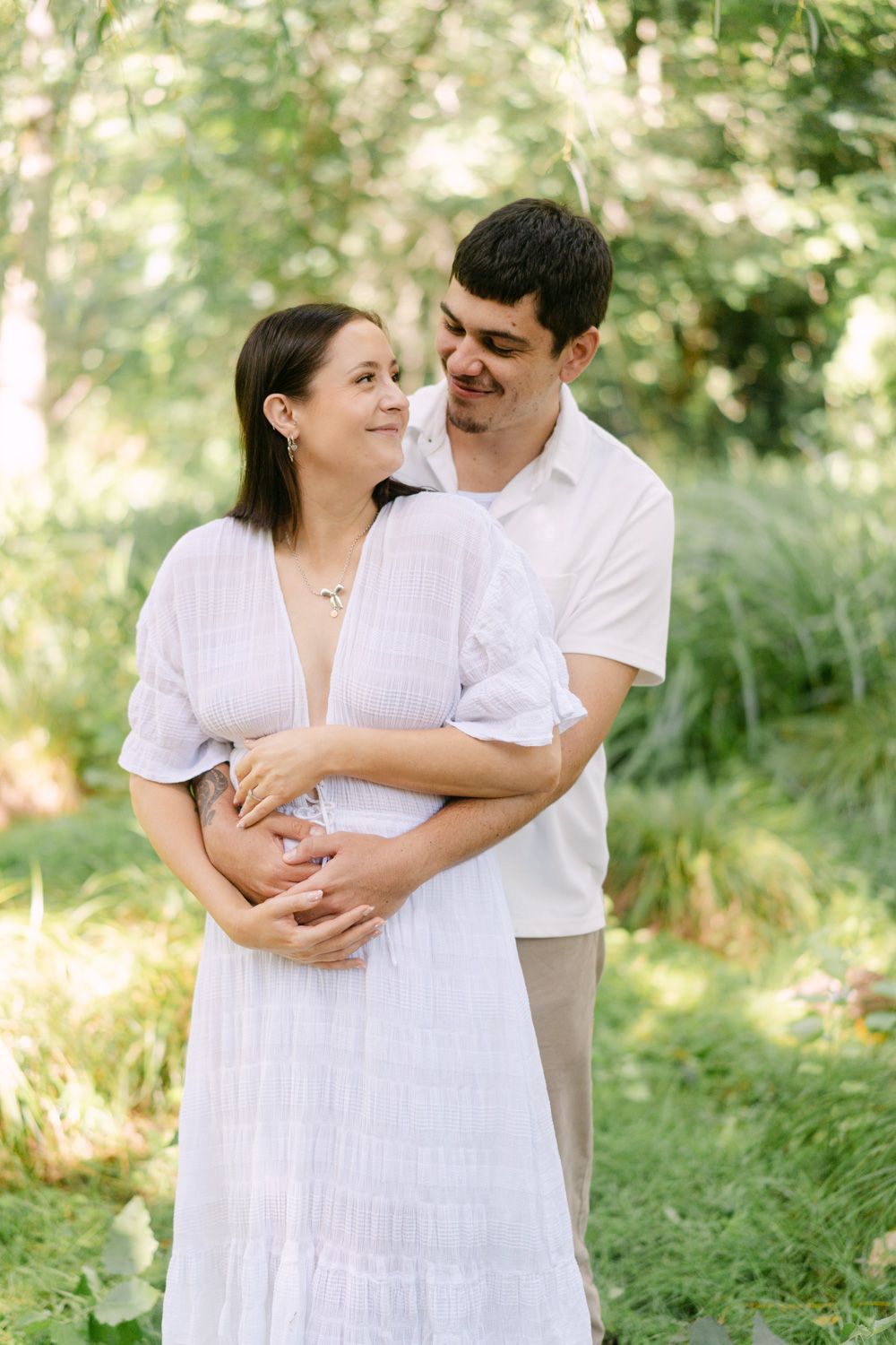 A couple embraces outdoors, the woman in a white dress cradling her baby bump, surrounded by lush green foliage.