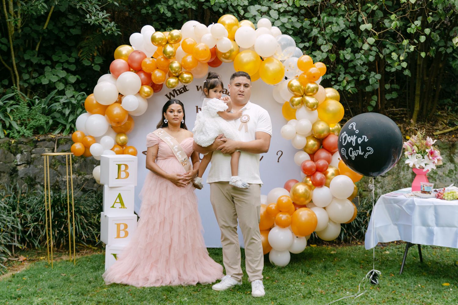 Family posing at a gender reveal party with colorful balloon arch, pink dress, and black gender reveal balloon.