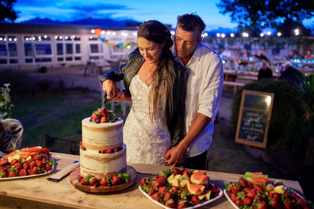 Bride and groom cutting a rustic naked wedding cake surrounded by fresh strawberries at an outdoor evening reception.