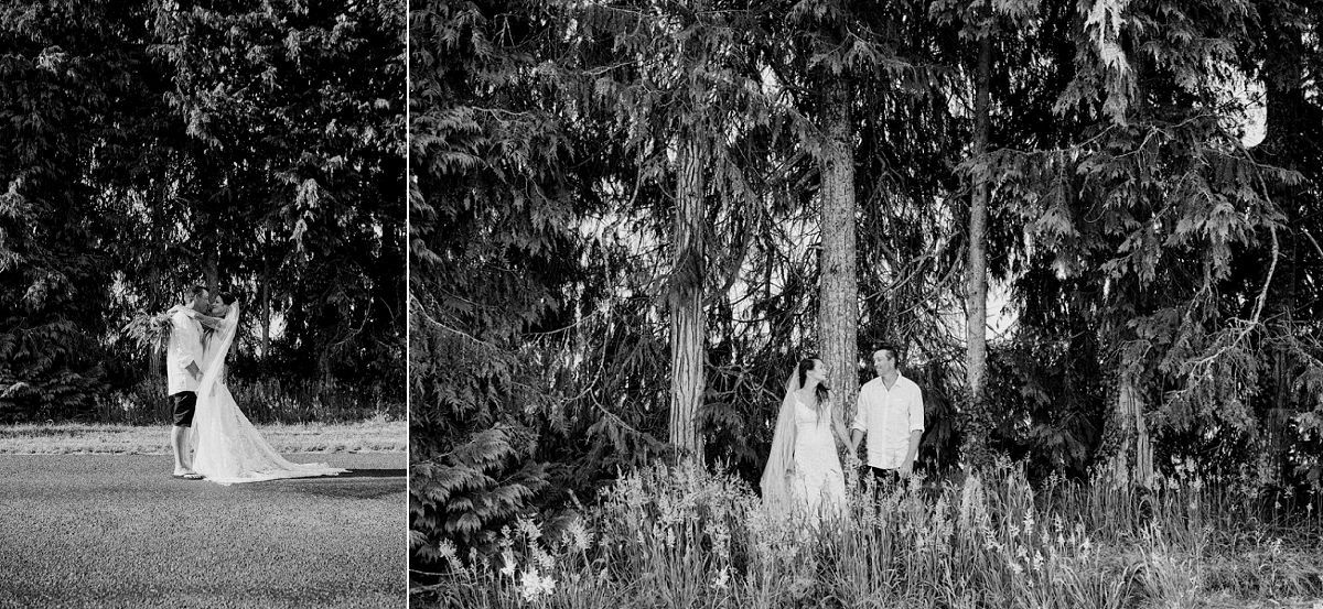 Black and white wedding photo of bride and groom posing near tall trees in a lush garden setting.