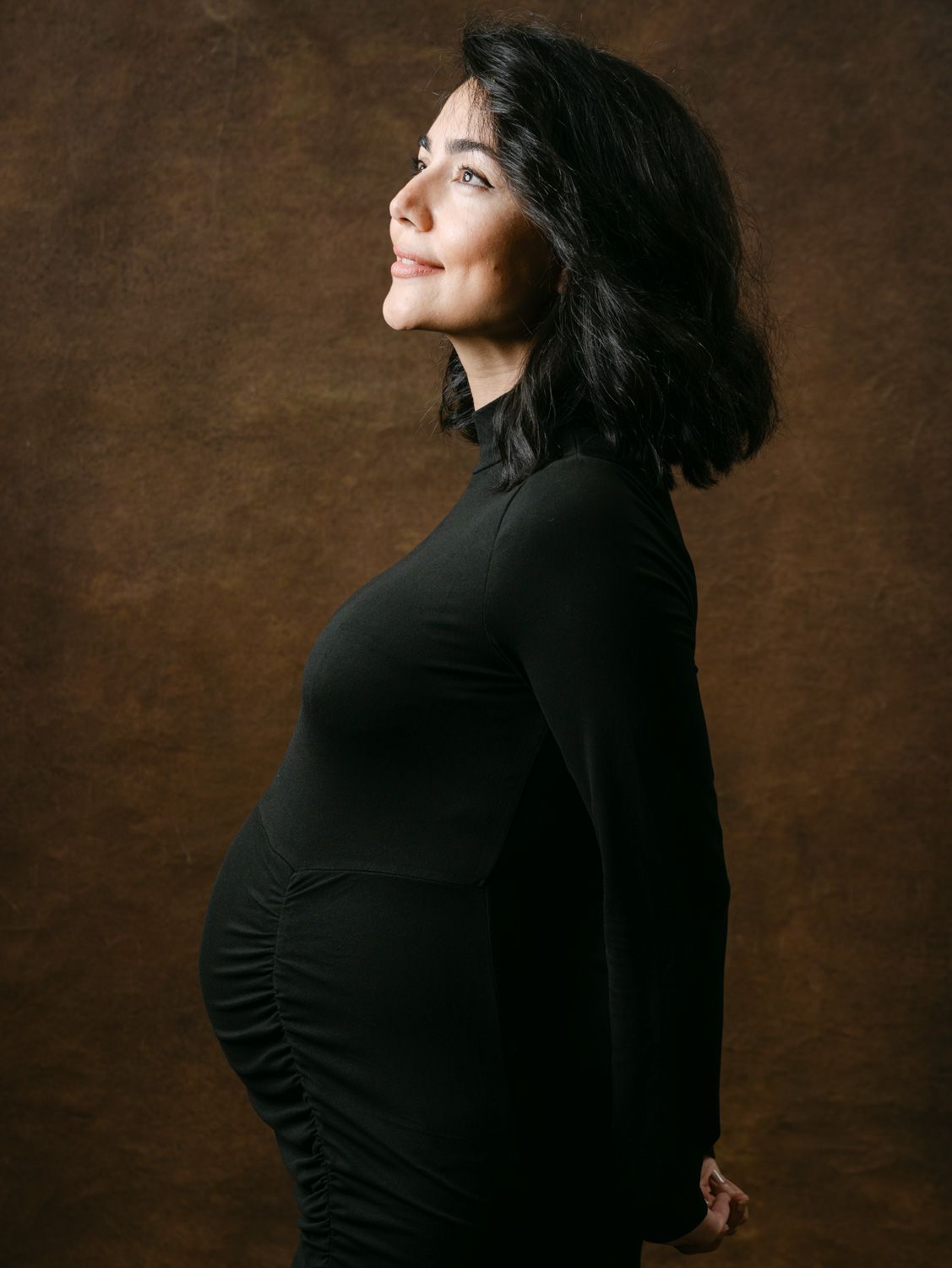 Pregnant woman in black turtleneck dress posing sideways, smiling, against brown studio background.
